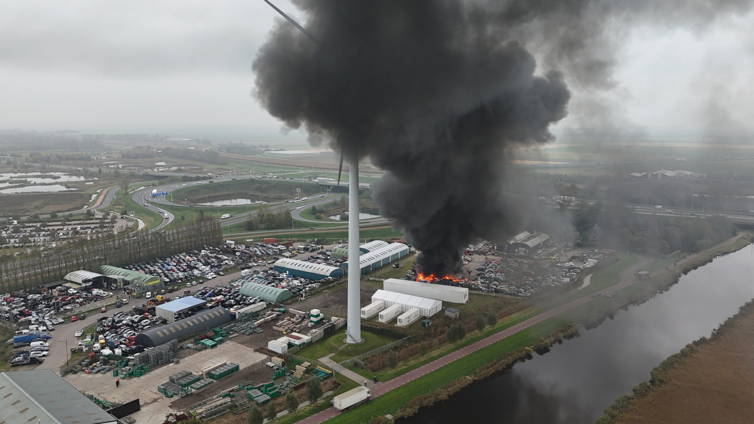 Luchtfoto van een industrieterrein met zwarte rook en vlammen opstijgend vanuit een gebouw, omgeven door auto's en een windmolen in de buurt.