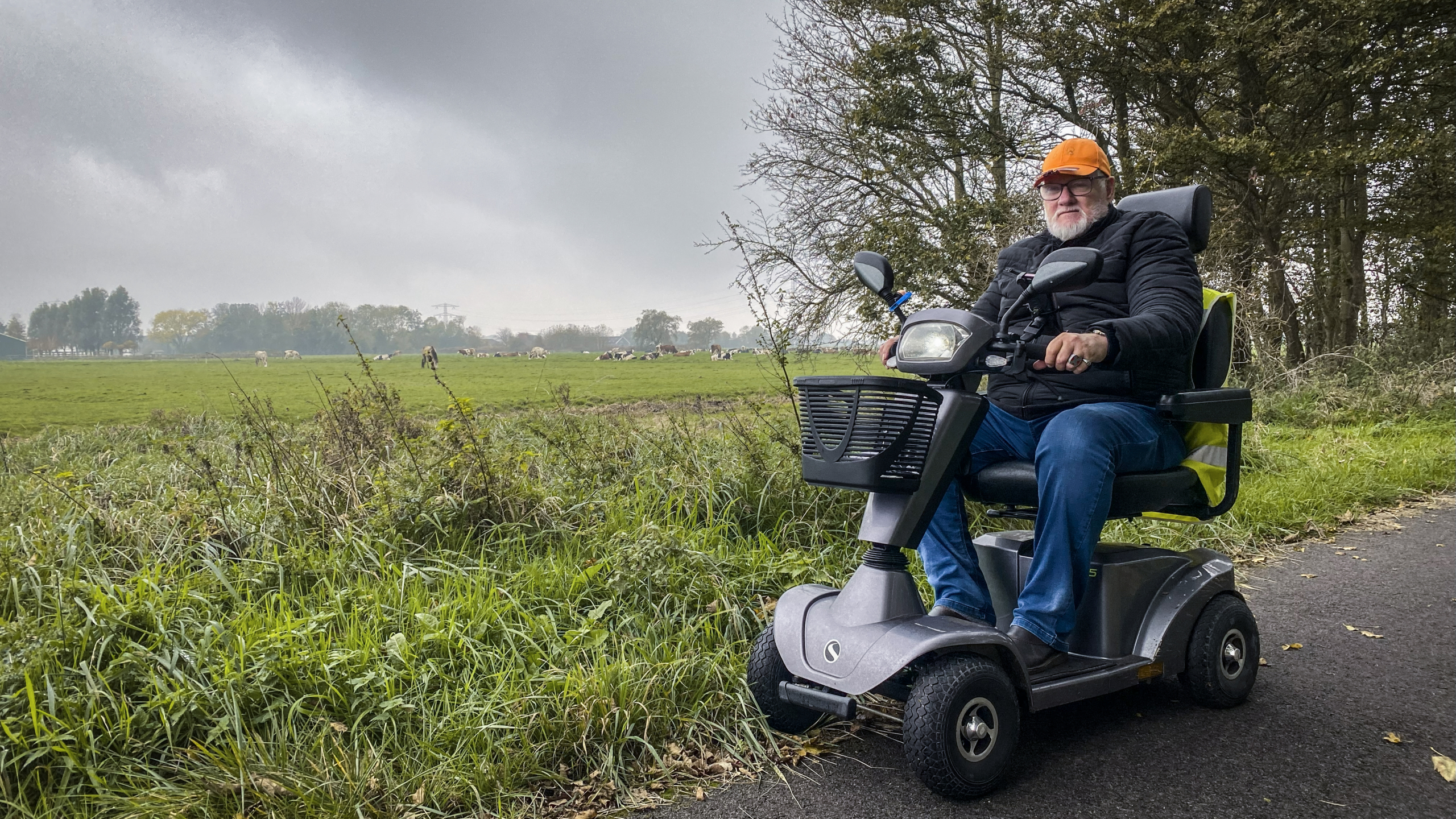 Man op een scootmobiel op een landweg naast grasvelden en bomen.