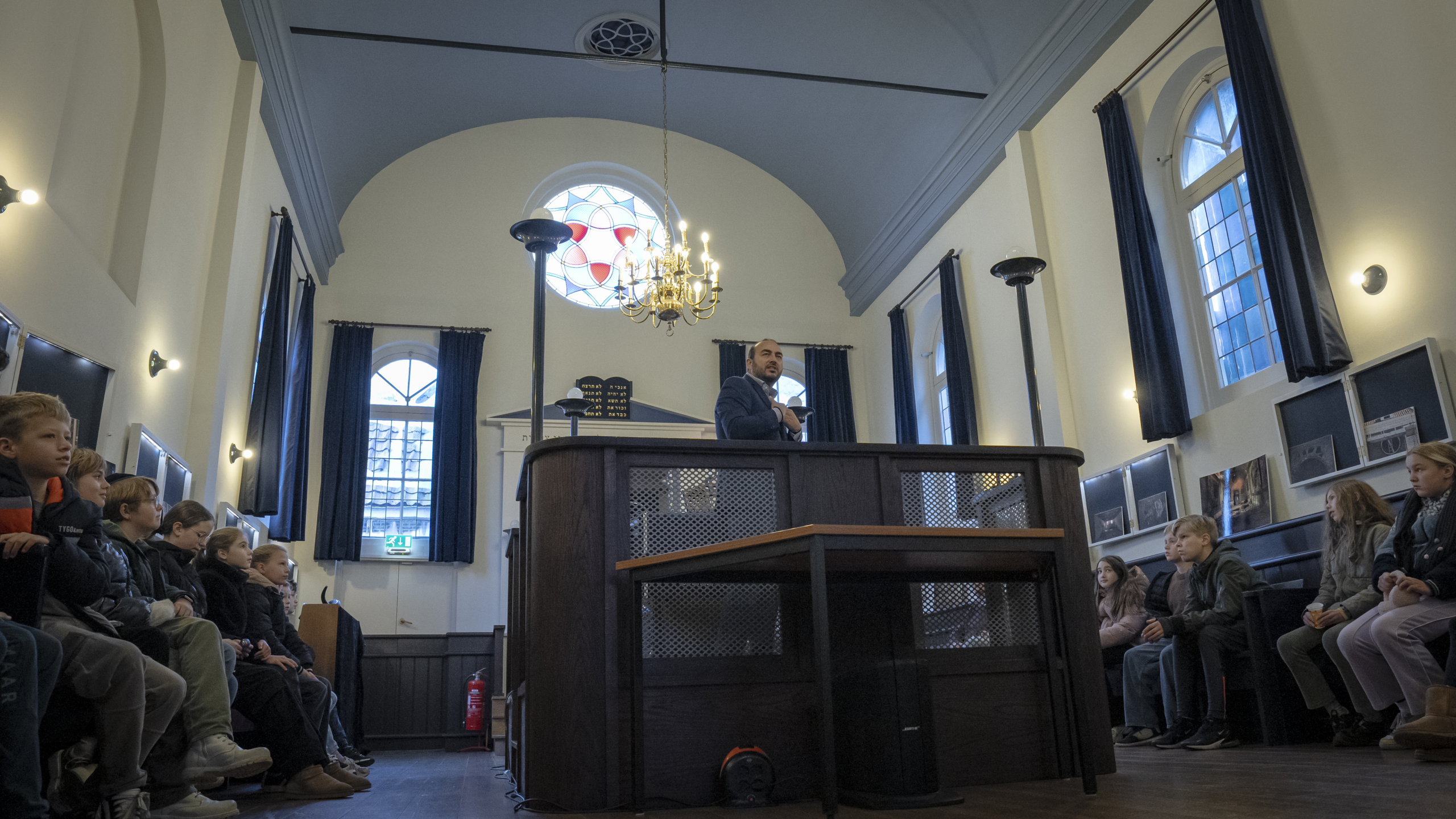 Mensen zitten in rijen in een zaal terwijl een spreker op een podium voor hen staat. De zaal heeft hoge ramen en een kroonluchter.