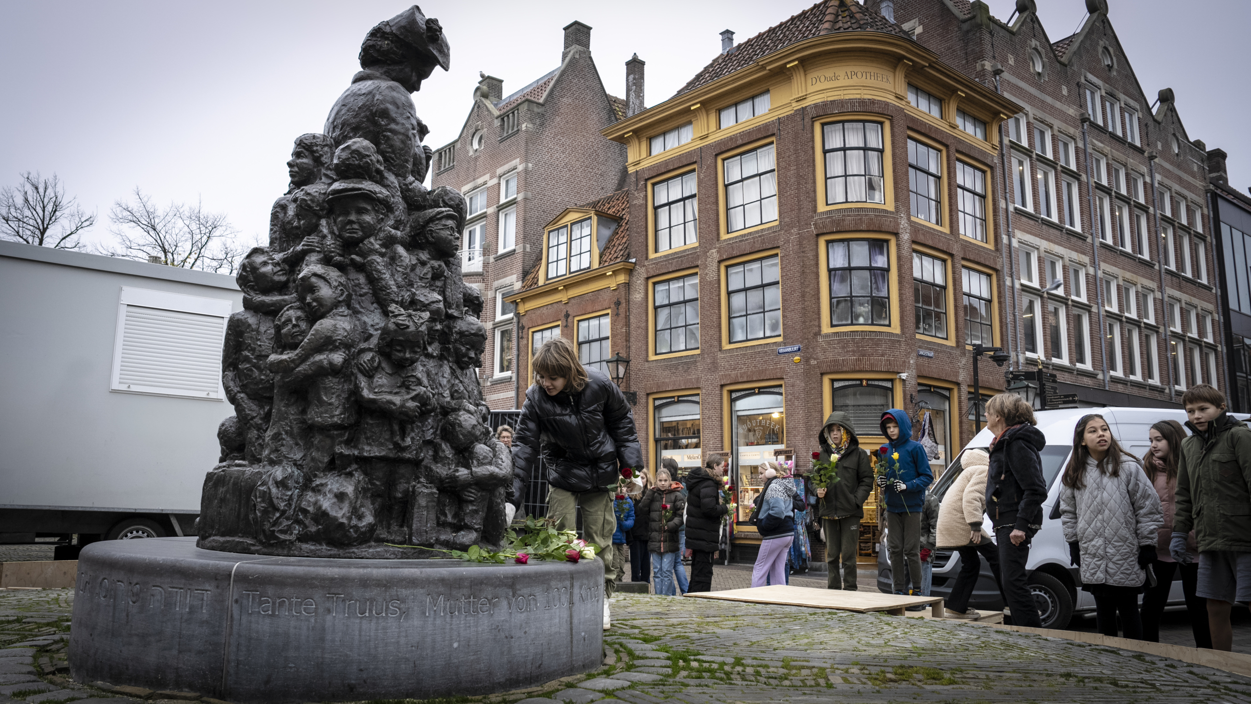 Beeld van een bronzen standbeeld met een vrouw en kinderen op straat, omgeven door mensen, met bloemen aan de voet. Op de achtergrond oude Nederlandse architectuur.