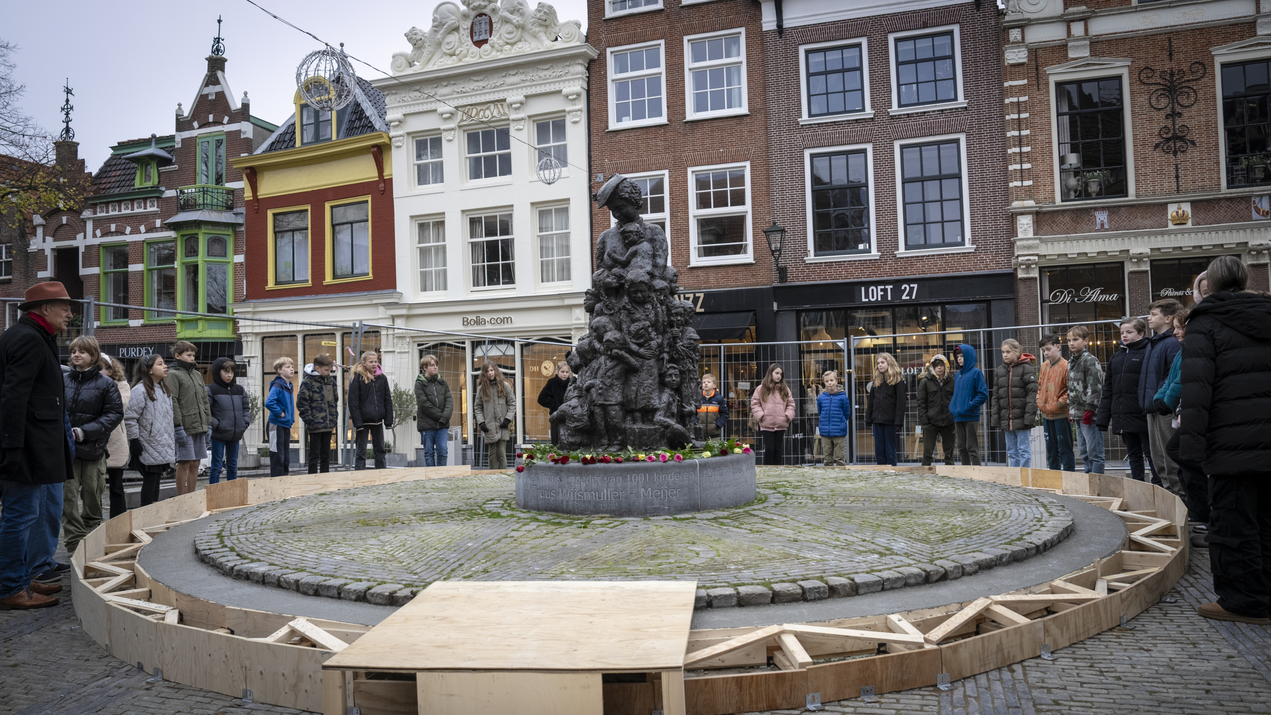 Een groep kinderen en een volwassene staan in een cirkel rond een bronzen standbeeld op een plein, omgeven door historische gebouwen.