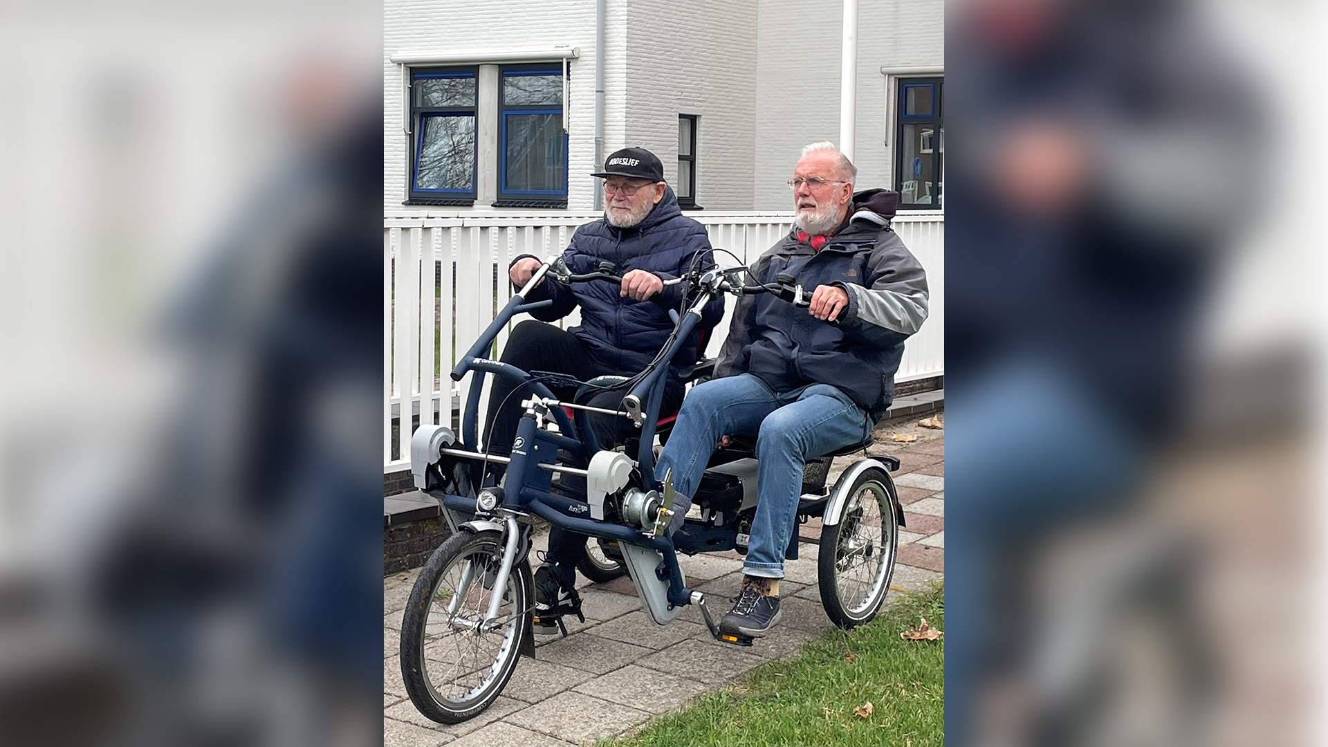 Twee mannen fietsen op een driewielige tandem voor een wit gebouw met blauwe ramen.