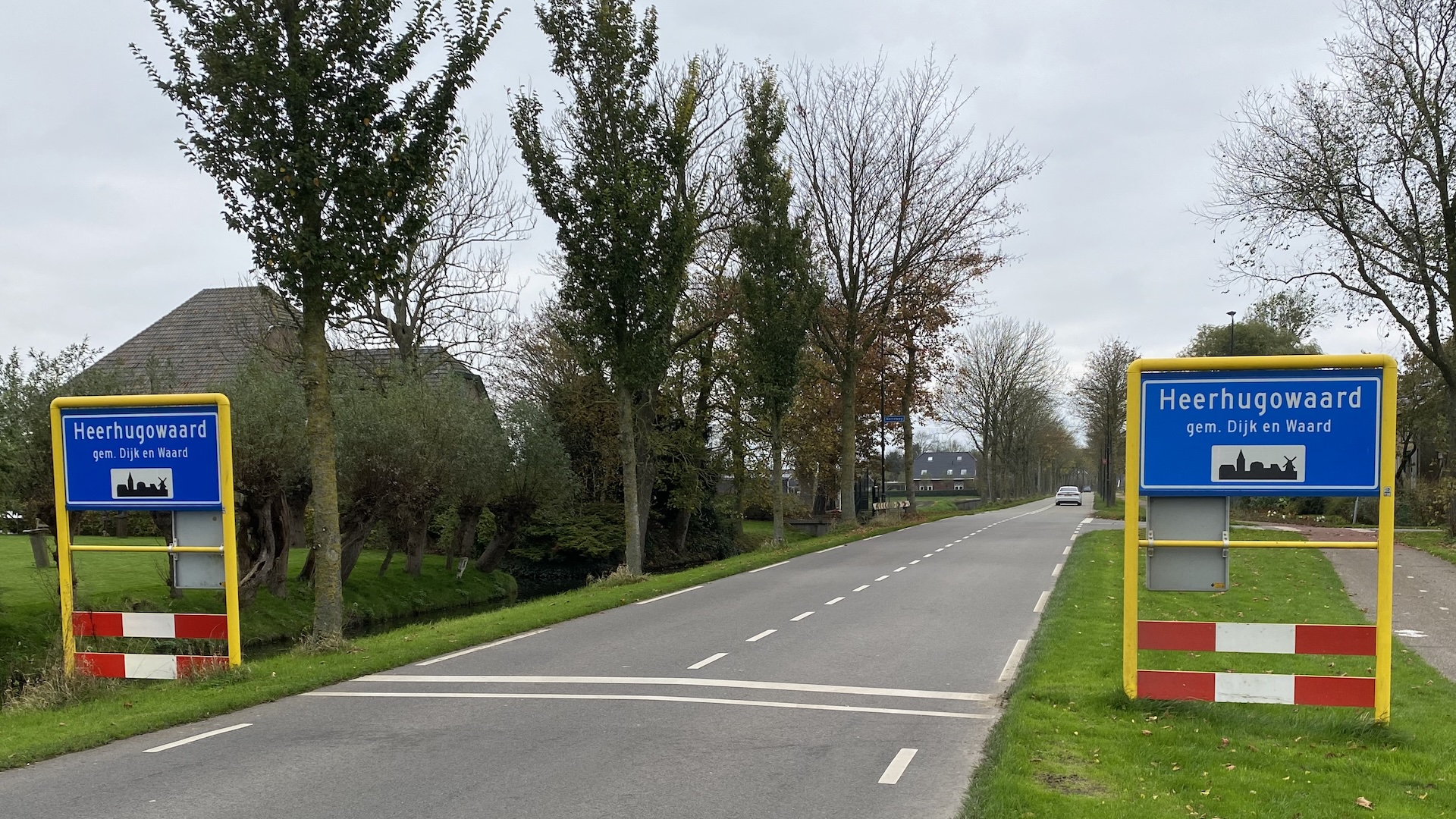 Twee borden met de tekst "Heerhugowaard, gem. Dijk en Waard" langs een weg, met bomen en een huis op de achtergrond.