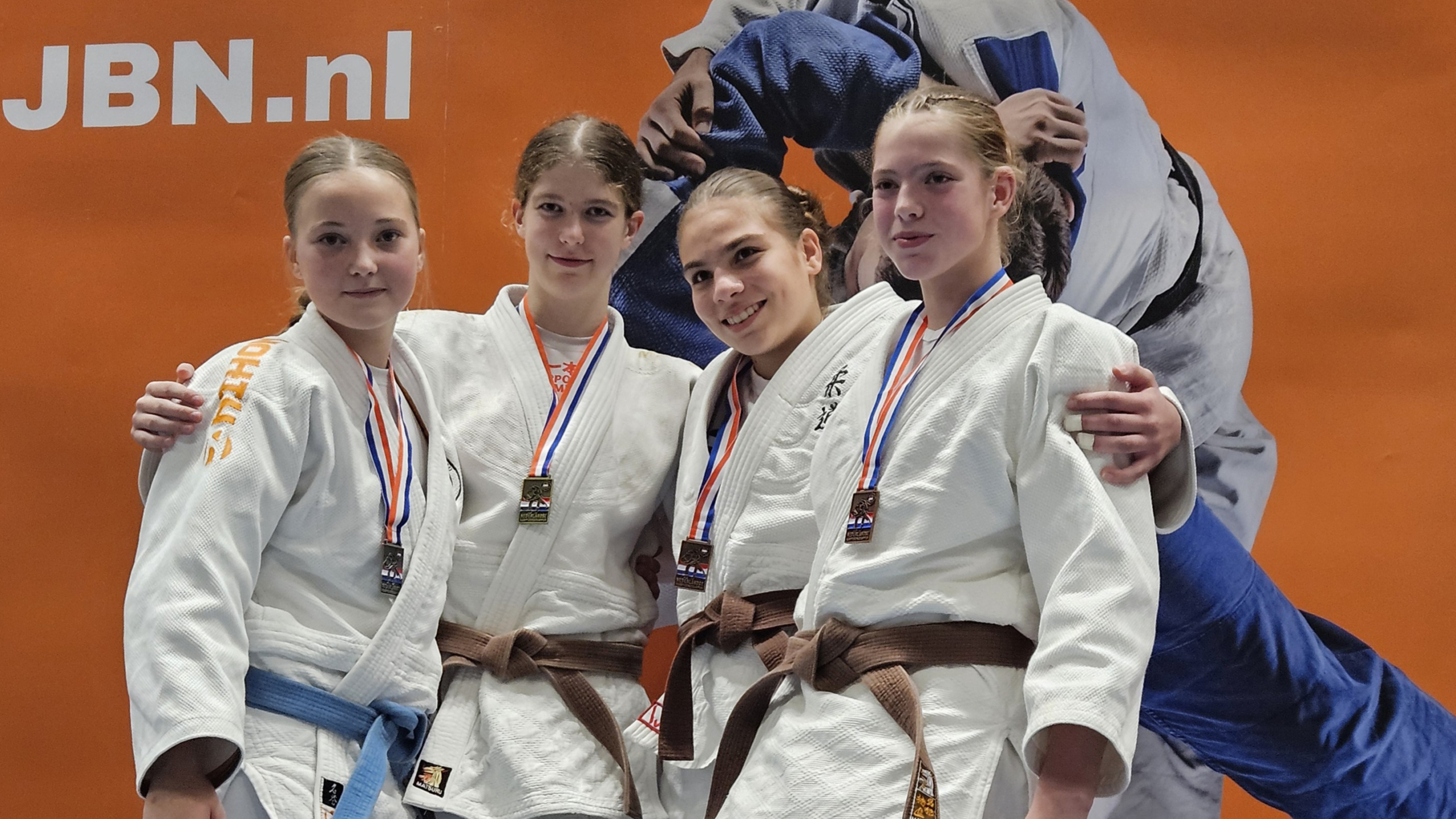 Vier jonge judoka's poseren met medailles voor een oranje achtergrond met het logo van JBN.nl.