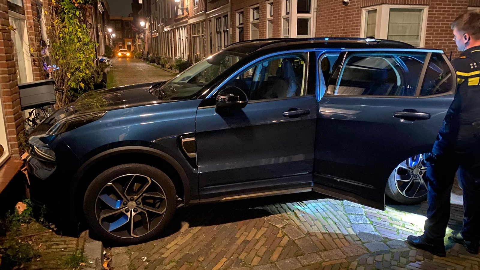 Auto geparkeerd op een smalle straat met een open deur, naast een bakstenen gebouw 's nachts; een persoon in een uniform staat naast de auto.