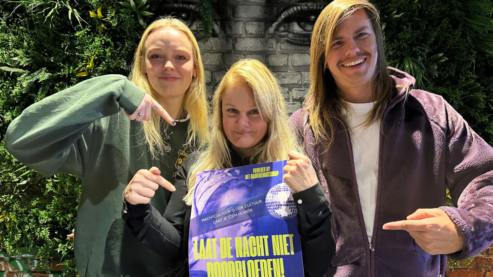 Twee vrouwen en een man poseren met een poster met de tekst "Laat de nacht niet doodblijven" voor een groene achtergrond.