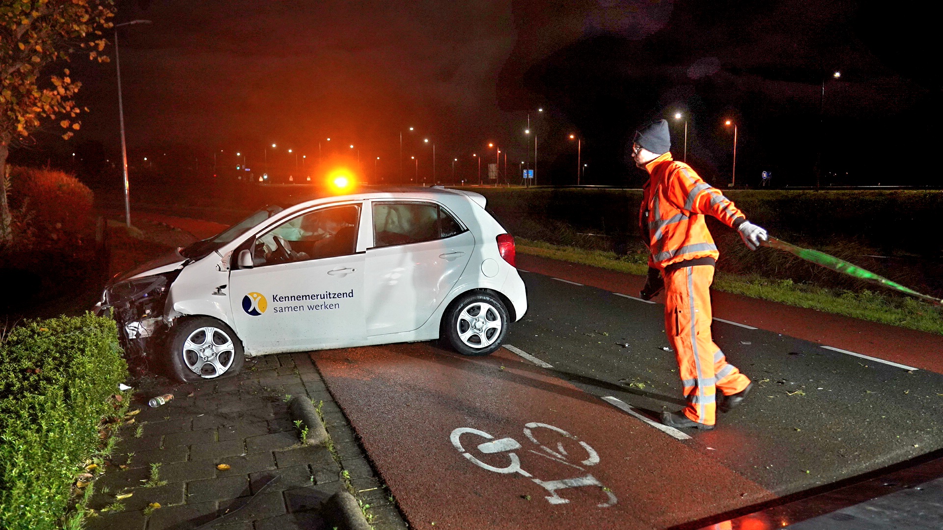 Een witte auto met logo van "Kennemeruitzend" is gecrasht tegen een heg. Een man in oranje reflecterend pak houdt een groen lint vast op een fietspad in het donker.