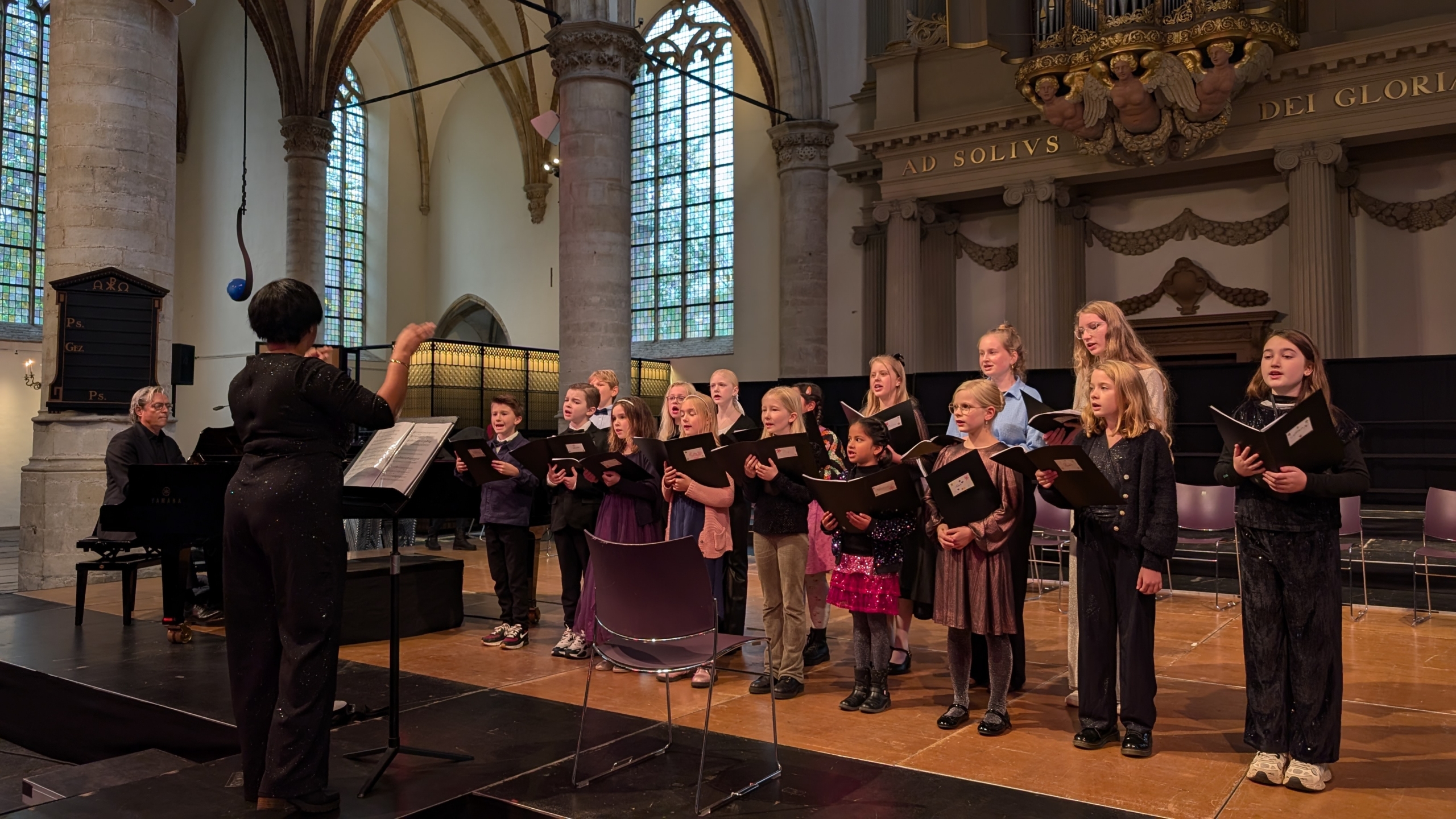 Kinderen zingen in een koor met een dirigent en pianist in een kerkachtige ruimte.