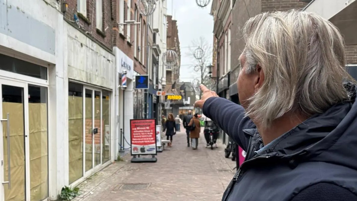 Een man met grijs haar wijst naar iets op een winkelstraat met mensen en een supermarkt bord in de verte.