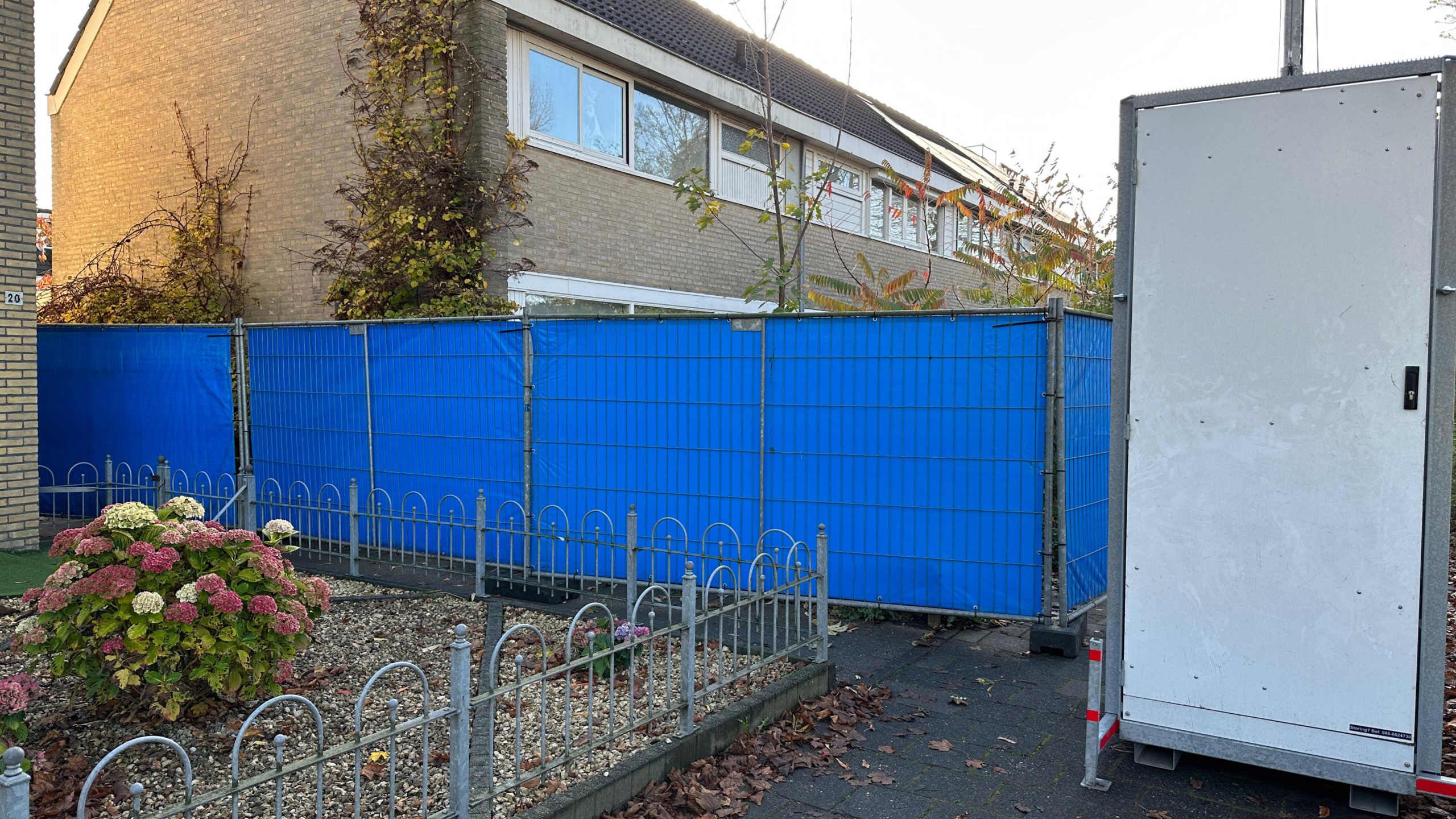 Blauwe bouwhekken naast een huis met een kleine tuin met hortensia's en een mobiel toilet op de stoep.