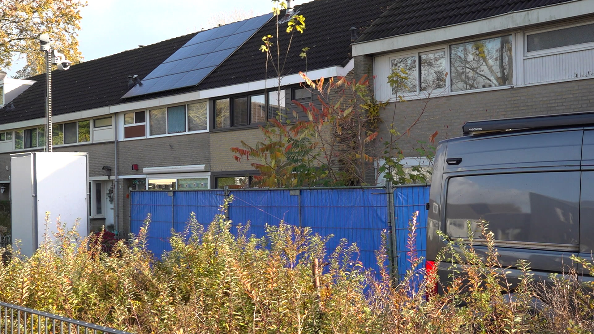 Rijtjeshuis met zonnepanelen op het dak, blauwe bouwhekken en een grijze bestelwagen op de voorgrond.