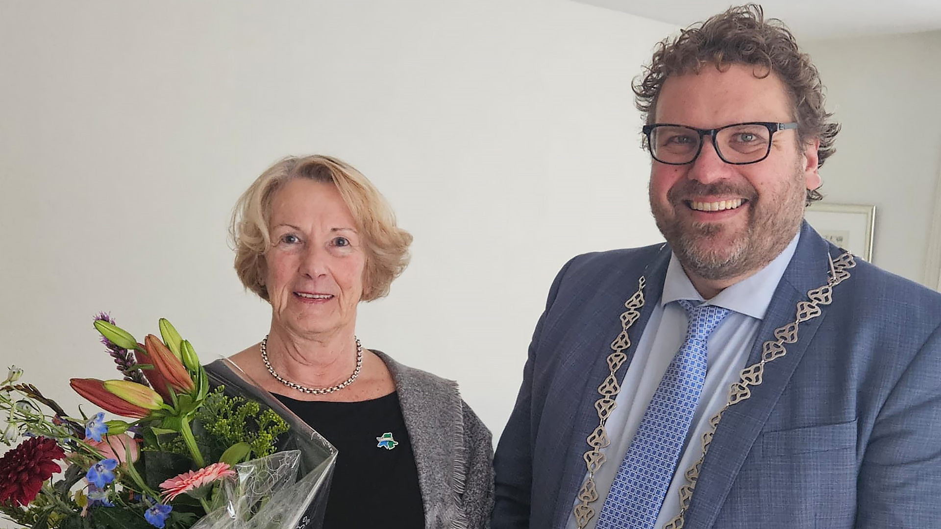 Een vrouw met een bloemenboeket en een man in een pak met ambtsketen glimlachen naar de camera.