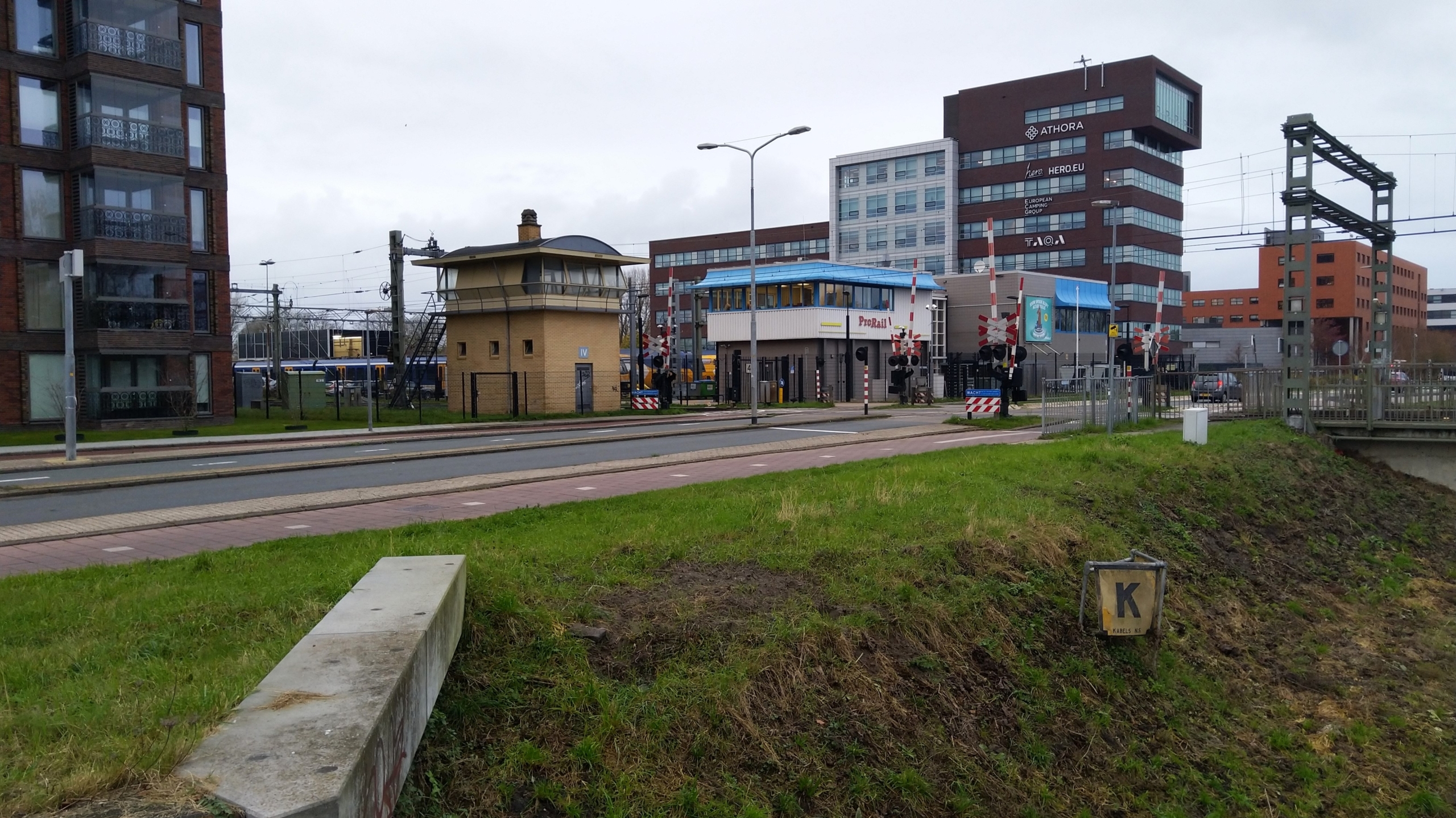 Een spoorwegovergang met nabijgelegen gebouwen, waaronder een hoge rode kantoorstructuur met de woorden "ATHORA" en "HERO.EU" erop.