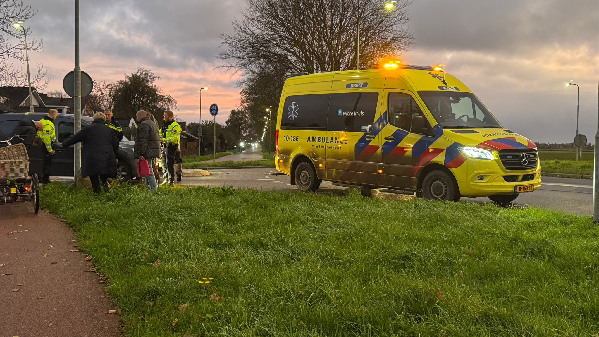 Gele ambulance met knipperende lichten op een kruispunt, omringd door mensen en politieagenten bij een grijskleurig voertuig in de schemering.