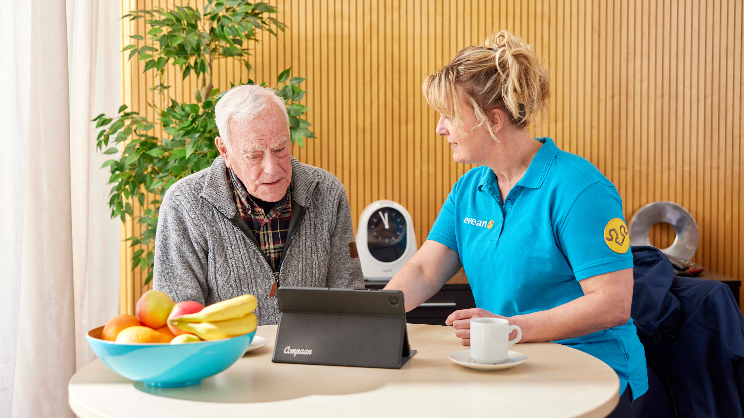 Een oudere man zit aan tafel en kijkt naar een tablet in gesprek met een vrouw in een blauw uniform.