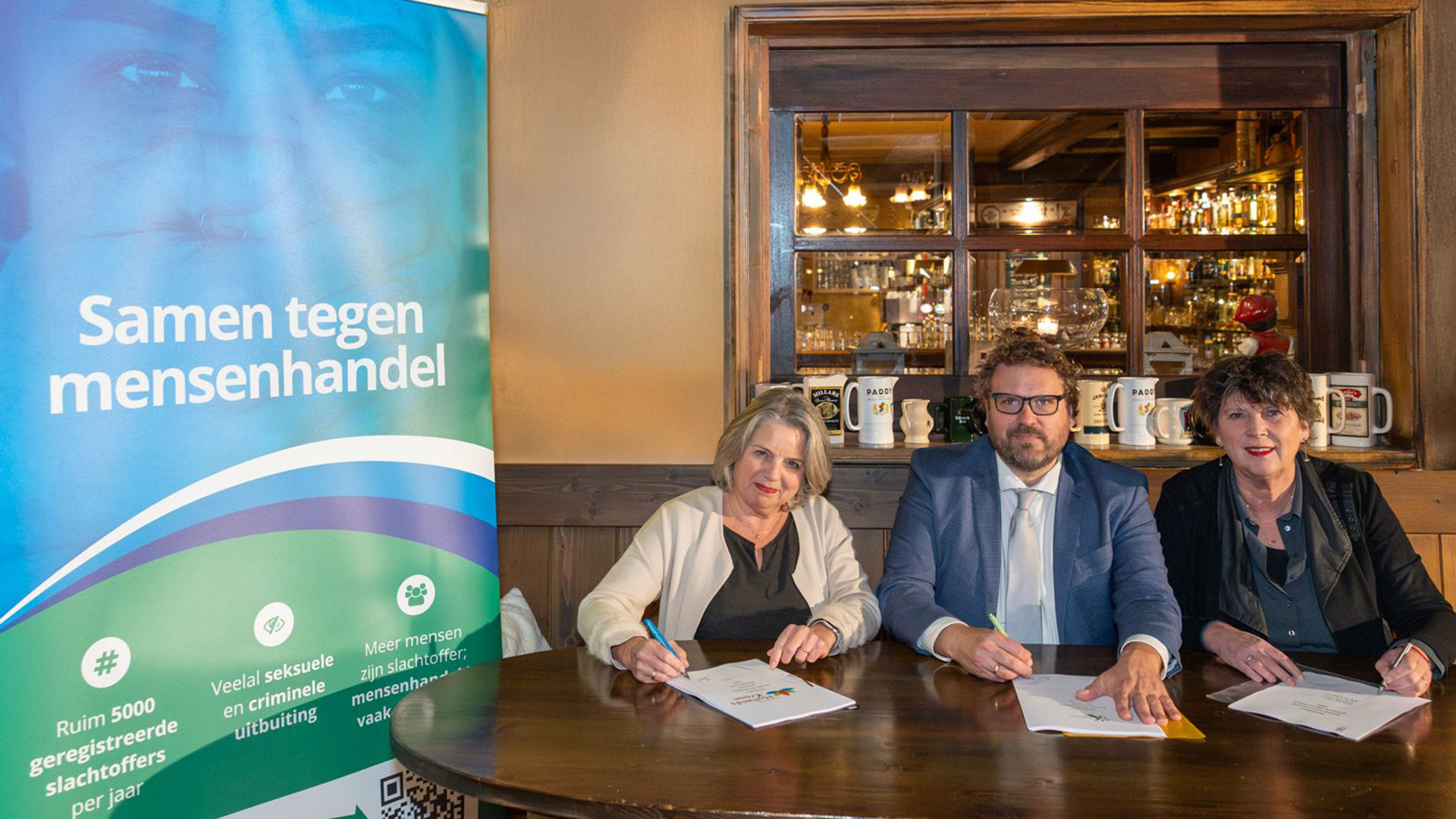 Drie mensen ondertekenen een document aan een tafel met een banner met de tekst "Samen tegen mensenhandel" op de achtergrond.