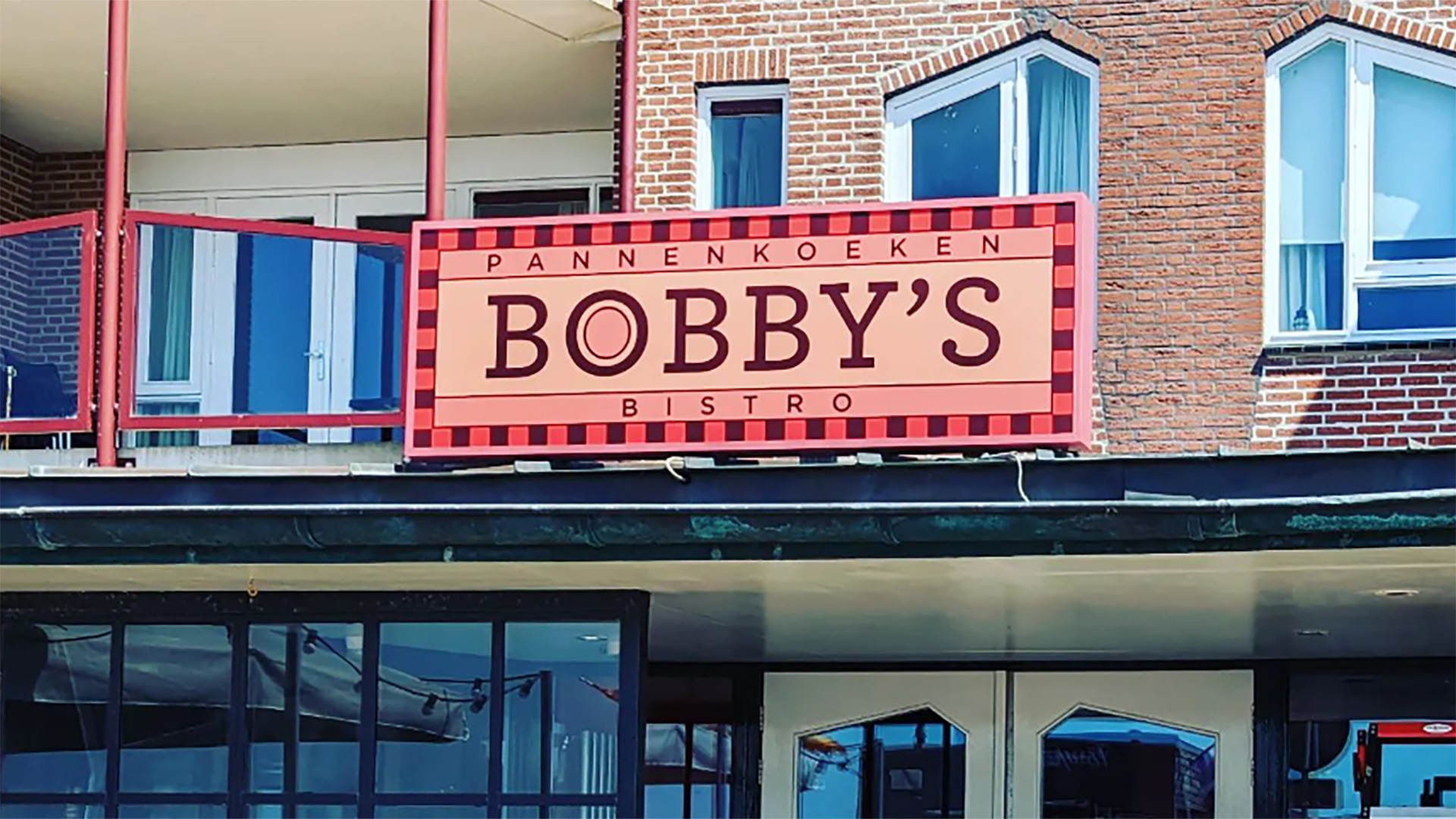 Uithangbord van "Pannenkoeken Bobby's Bistro" boven de ingang van een gebouw met bakstenen muur en balkon.