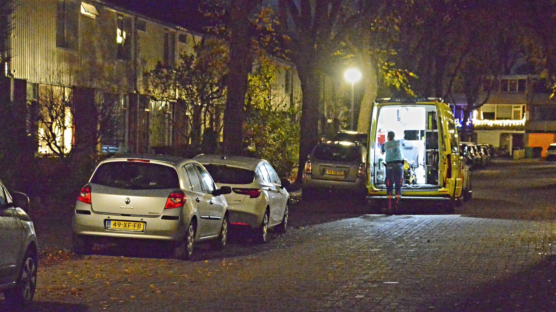 Een ambulance staat geparkeerd in een woonwijk bij nacht, met geopende achterdeuren en verlichting aan; een persoon staat bij de ambulance.