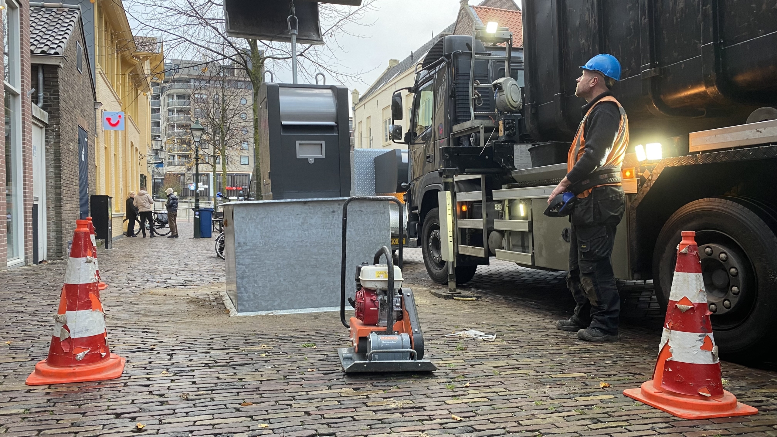 Bouwplaats met een vrachtwagen en arbeider met helm, omgeven door oranje verkeerskegels.