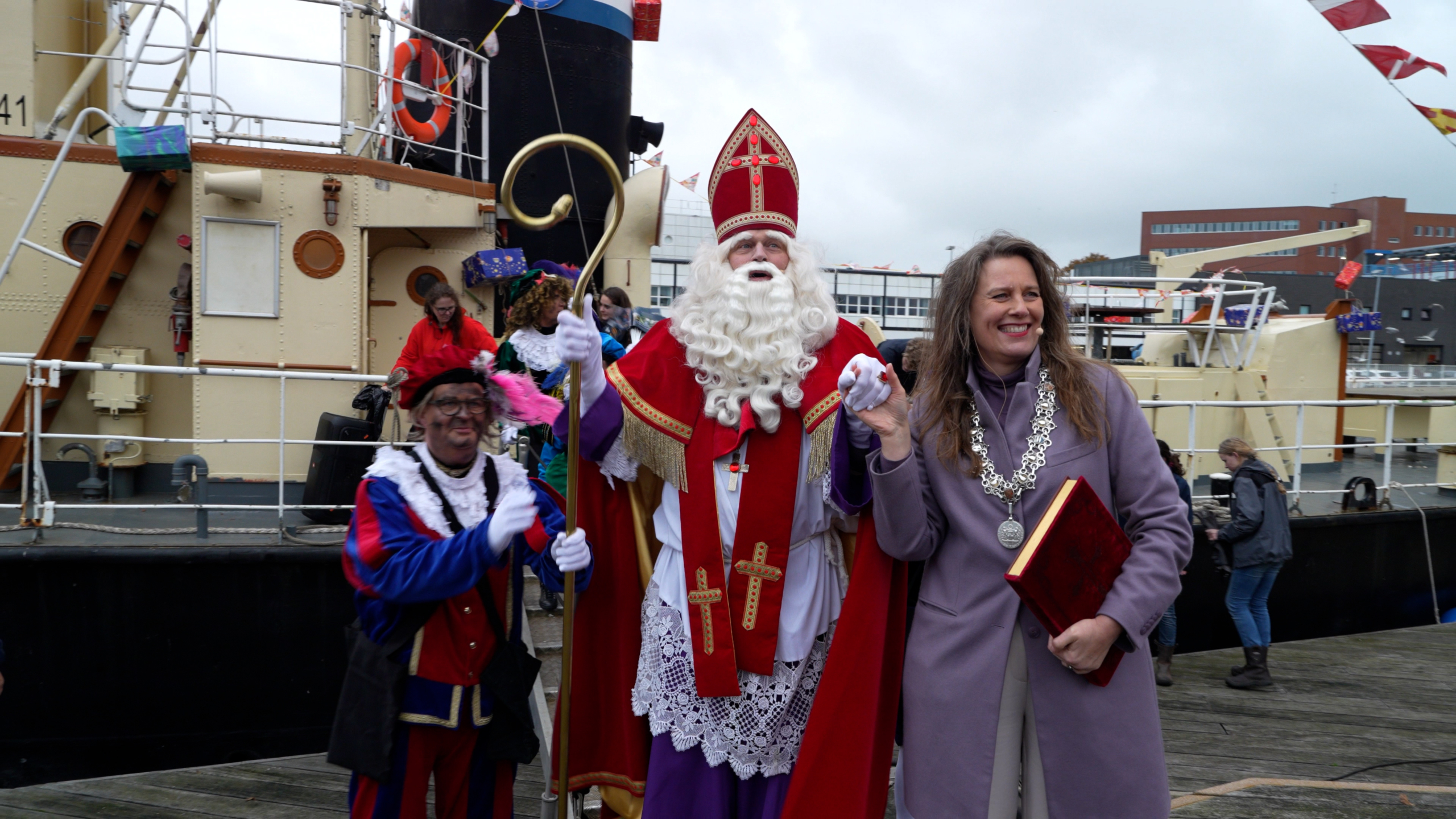 Sinterklaas en Pieten arriveren op een boot, begroet door een lachende vrouw met een ambtsketting.