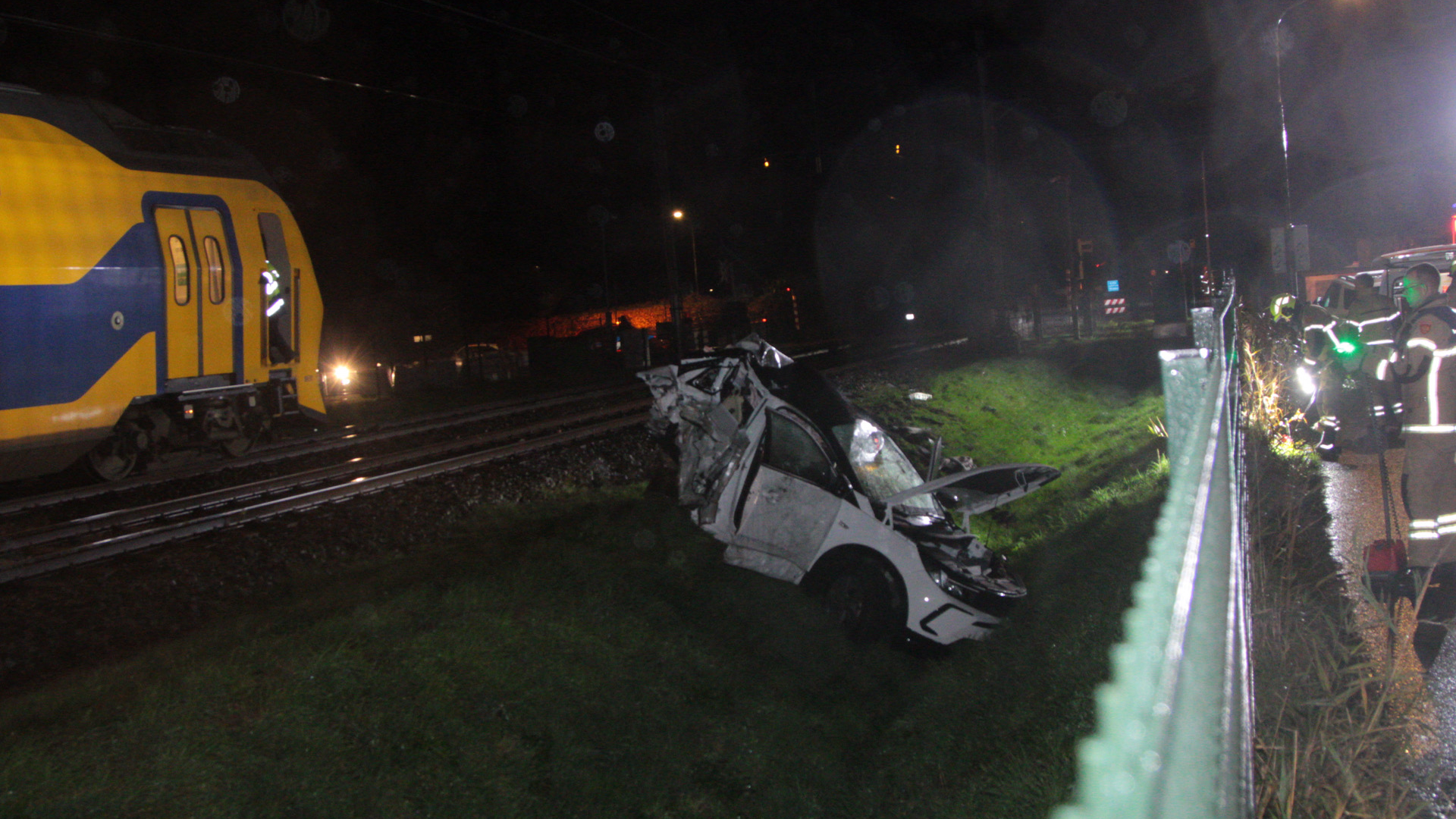 Een trein naast een zwaar beschadigde auto langs het spoor, met hulpverleners ter plaatse in de nacht.