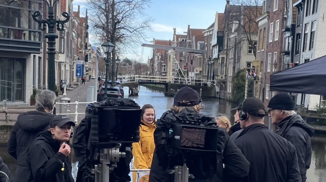 Groep mensen verzameld in een historische stad bij een gracht met op de voorgrond een vlag met illustratie en een zwarte tent.