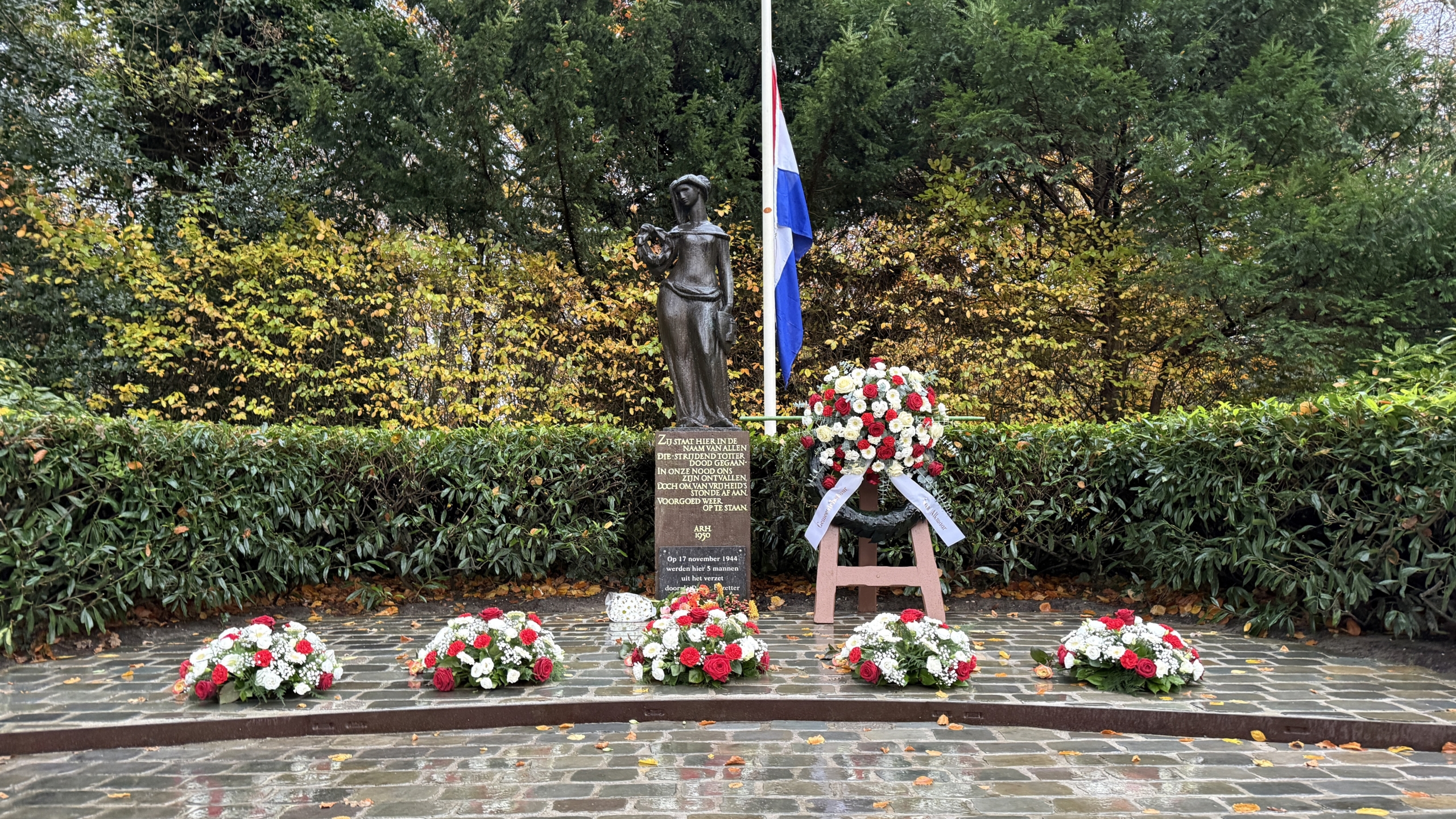 Een oorlogsmonument met een beeld, een Nederlandse vlag halfstok, en kransen van bloemen op een regenachtige dag.