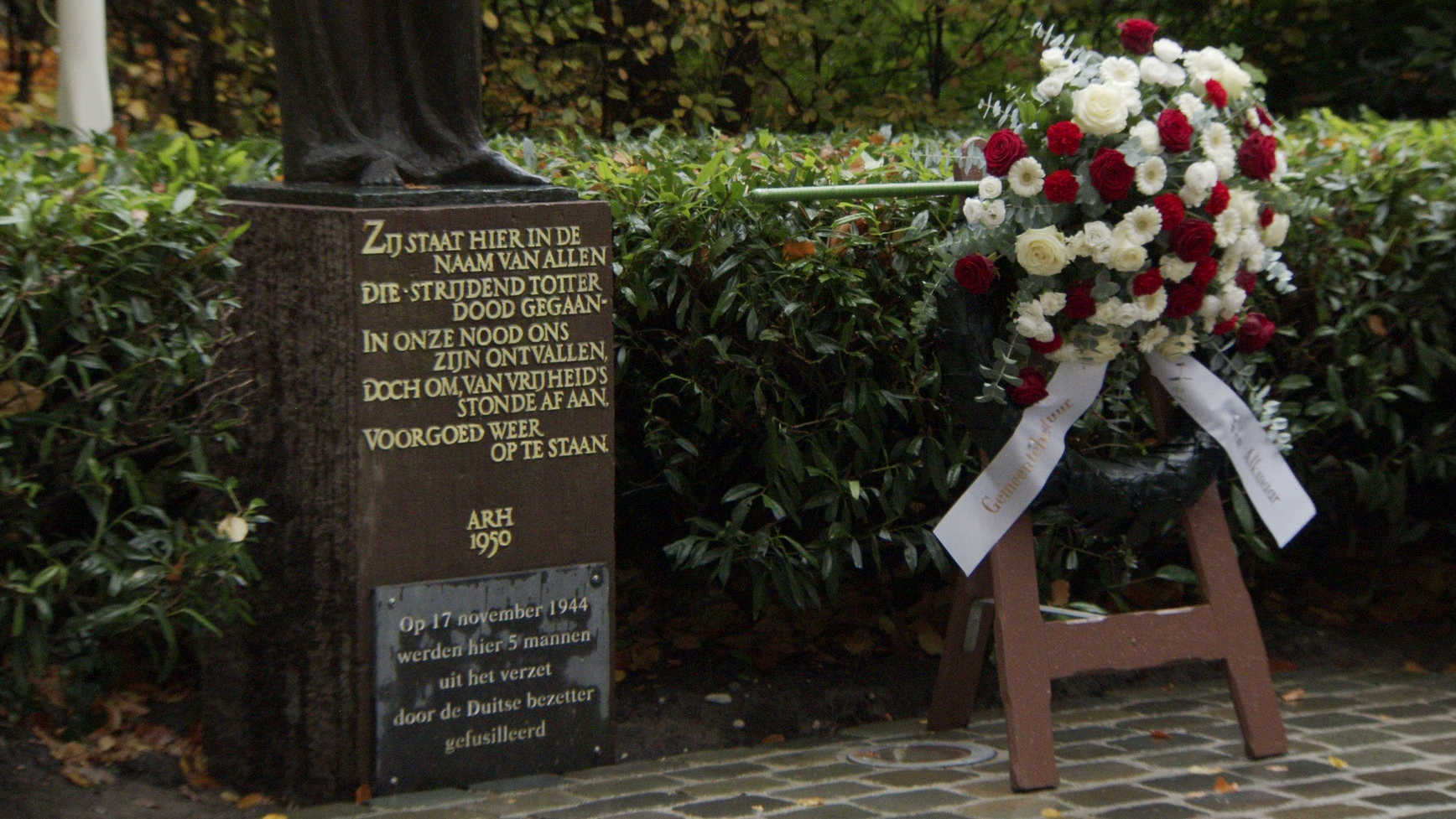 Een gedenksteen met een tekst ter nagedachtenis aan verzetsstrijders en een bloemstuk met rode en witte bloemen.