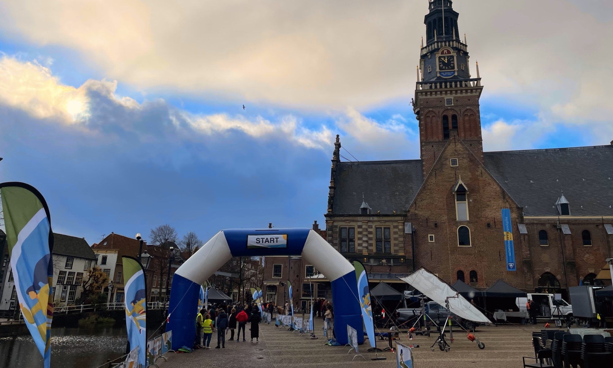 Een startboog met vlaggen op een plein bij een historisch gebouw met klokkentoren in Alkmaar.