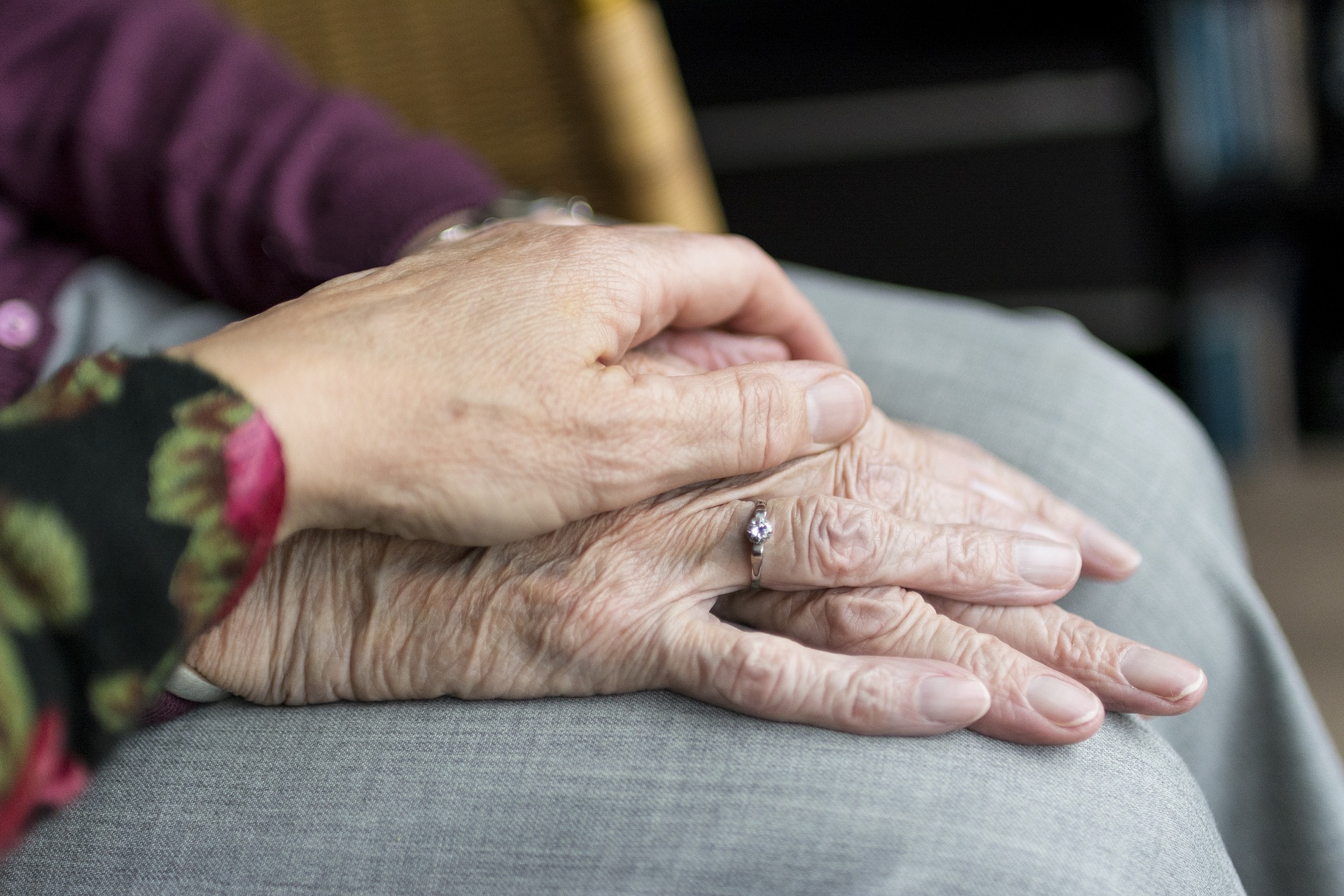 Twee handen die elkaar vasthouden, een oudere hand met een ring en een jongere hand erbovenop, op een schoot met grijze stof.