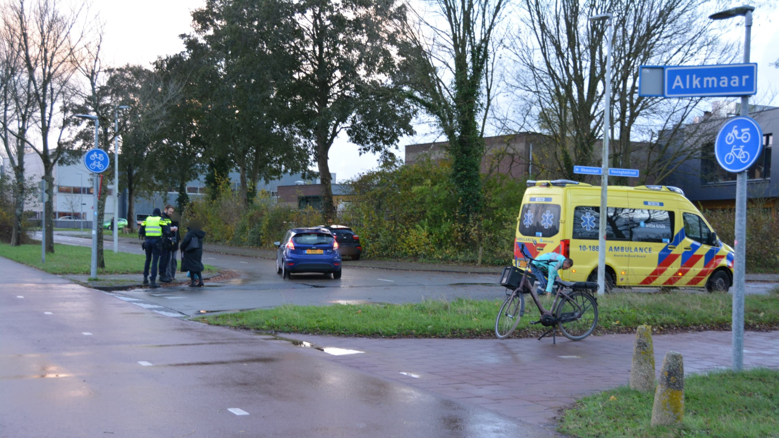 Fiets met ambulance en politieagenten bij een weg met een bord richting Alkmaar.