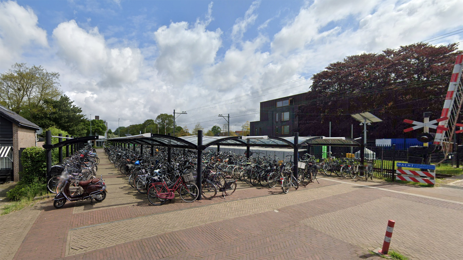 Fietsenstalling met veel geparkeerde fietsen onder een overkapping, naast een spoorwegovergang.