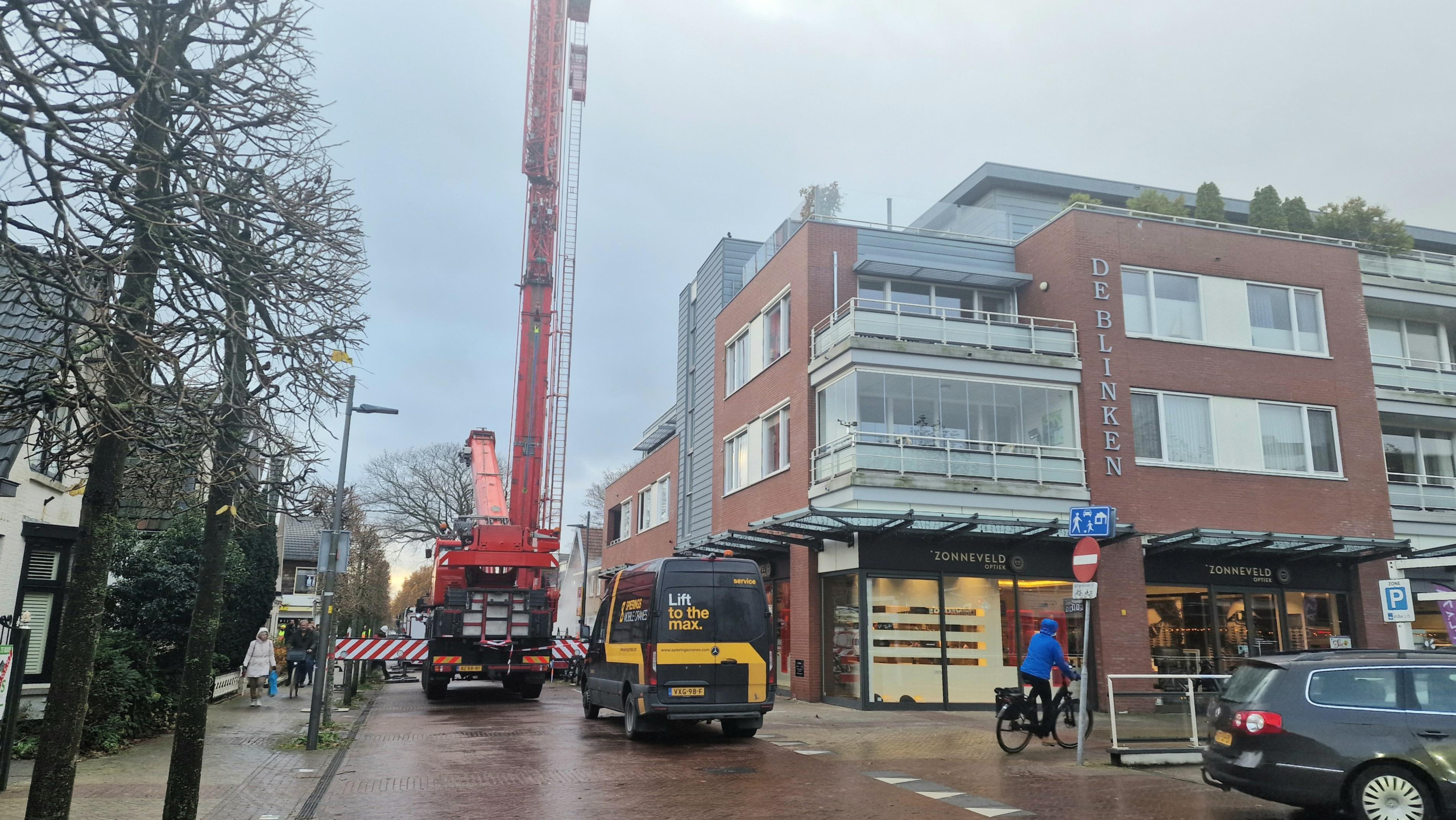 Een straatbeeld met een rode kraan naast een gebouw genaamd "De Blinken", een bestelwagen met opschrift "Lift to the max", en een fietser aan de rechterzijde.