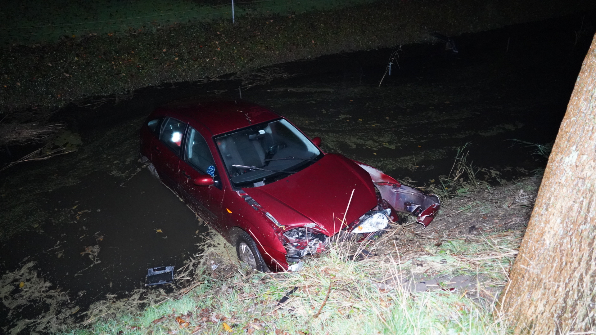 Rode auto half ondergedompeld in een sloot, met beschadigde voorkant, nachtelijk tafereel.