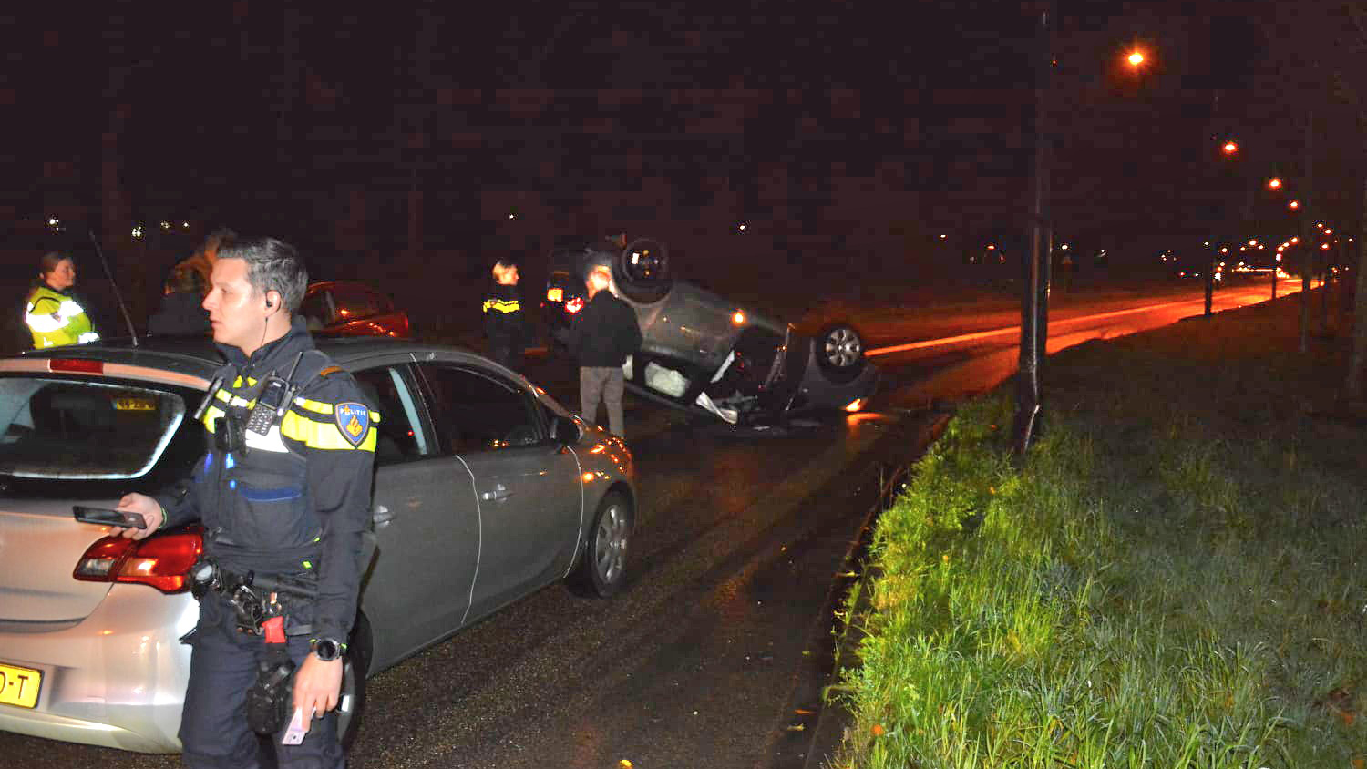 Auto-ongeluk op een natte weg in het donker, met politieagenten die de scène onderzoeken.