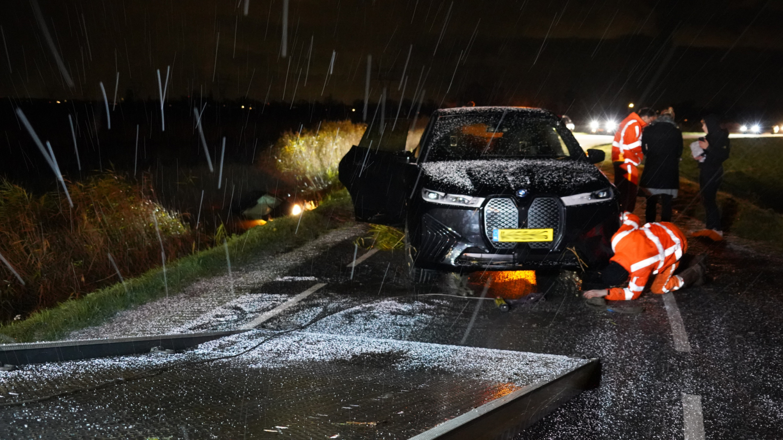 Auto langs de weg met schade, omringd door mensen in reflecterende kleding, 's nachts in de regen.