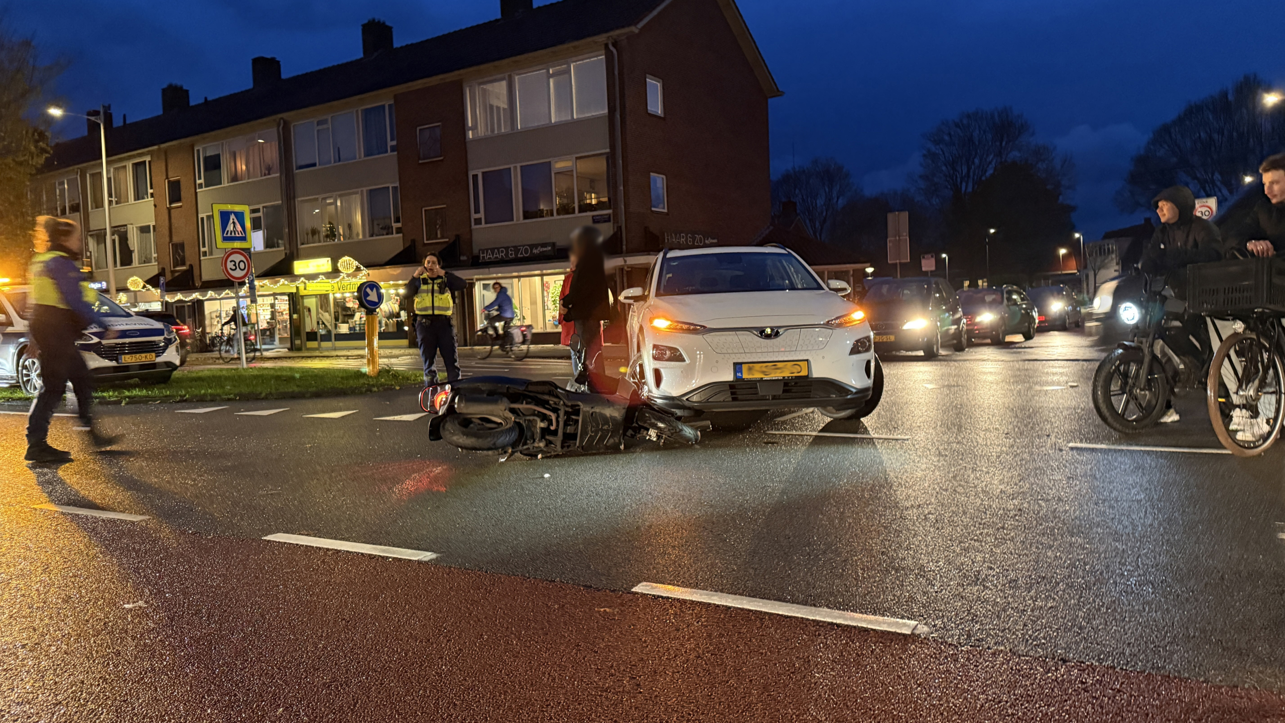 Een auto is betrokken bij een aanrijding met een omgevallen scooter op een drukke weg, omgeven door mensen en verkeer; politieagenten zijn ter plaatse.