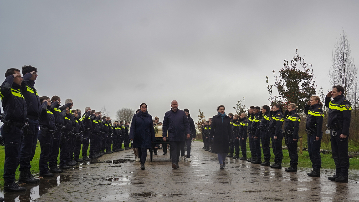 Mensen in uniform staan in twee rijen en brengen een saluut terwijl anderen een kist naar voren dragen onder een grijze, bewolkte lucht.