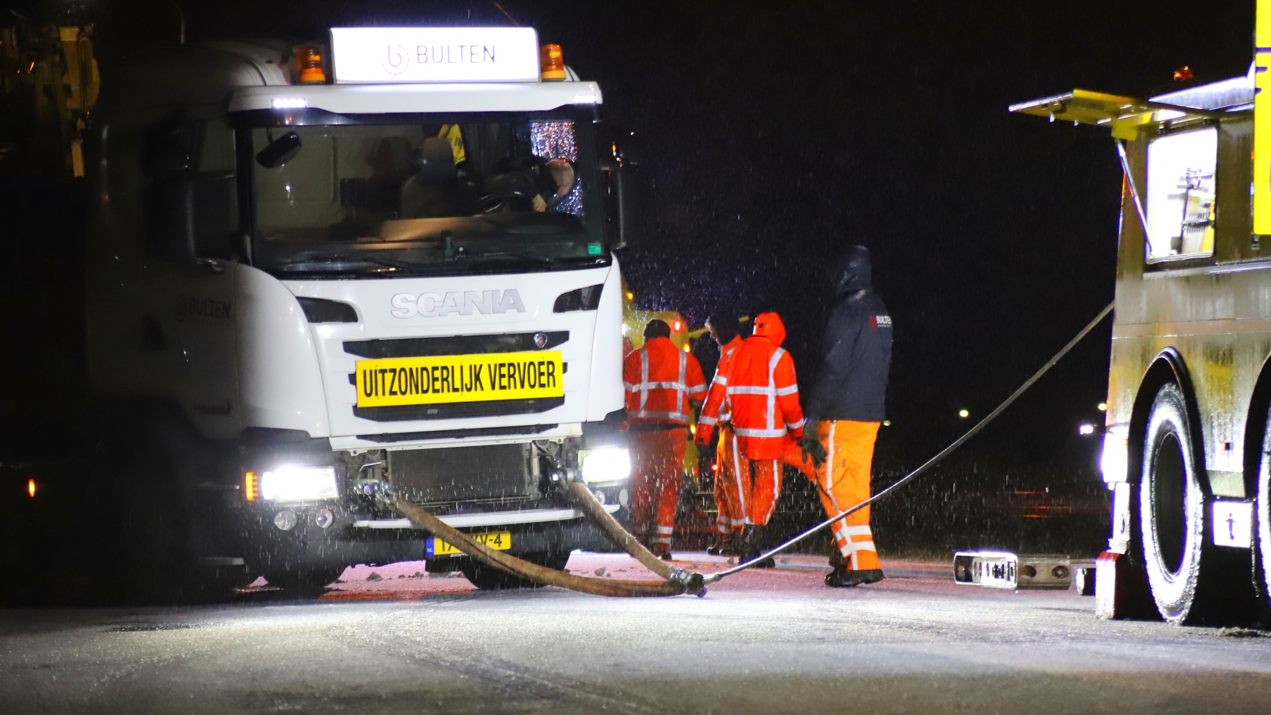 Vrachtwagen met bord "UITZONDERLIJK VERVOER" op een nat wegdek 's nachts, omringd door mannen in oranje veiligheidskleding.