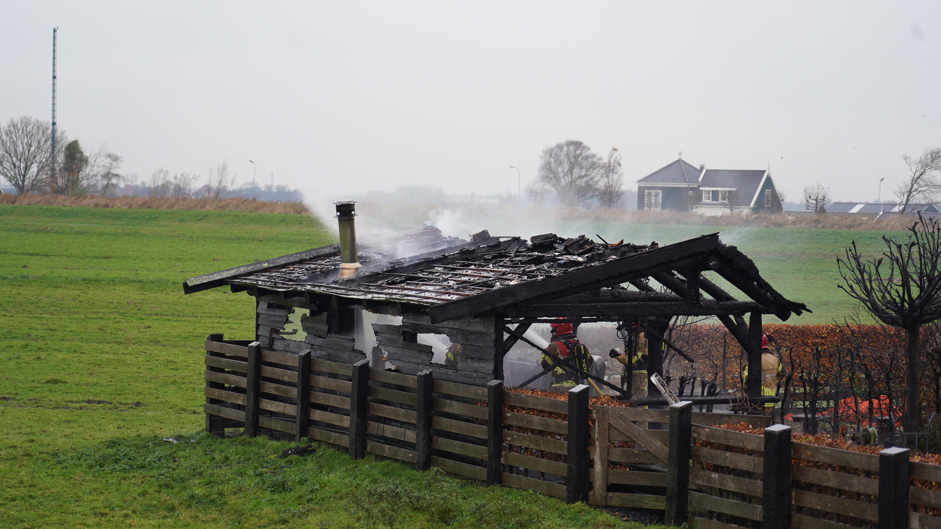 Verbrand bijgebouw met bluswerkzaamheden van brandweerlieden in een weiland.