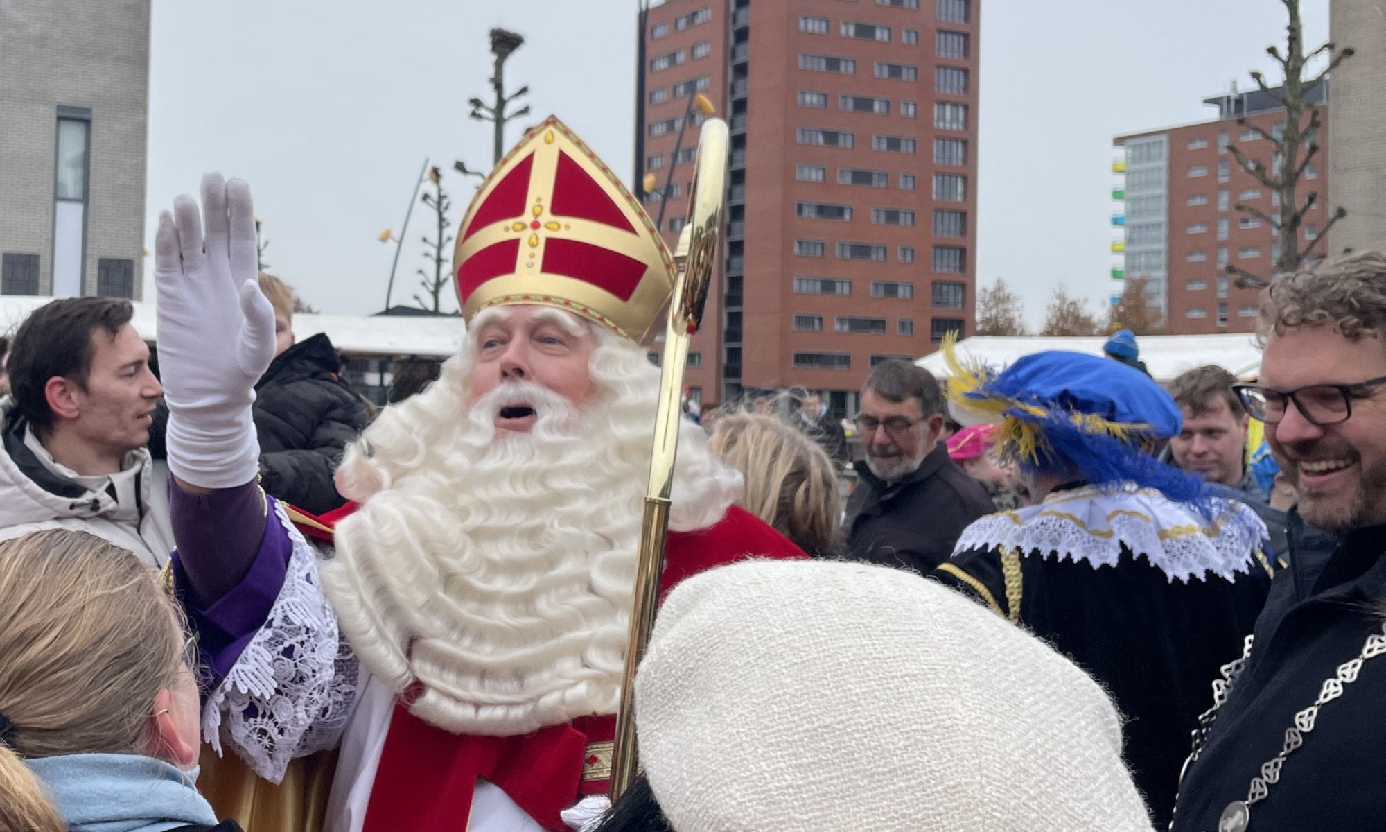 Sinterklaas begroet mensen in een menigte, bijgestaan door een Piet en omringd door toeschouwers in een stadsomgeving.