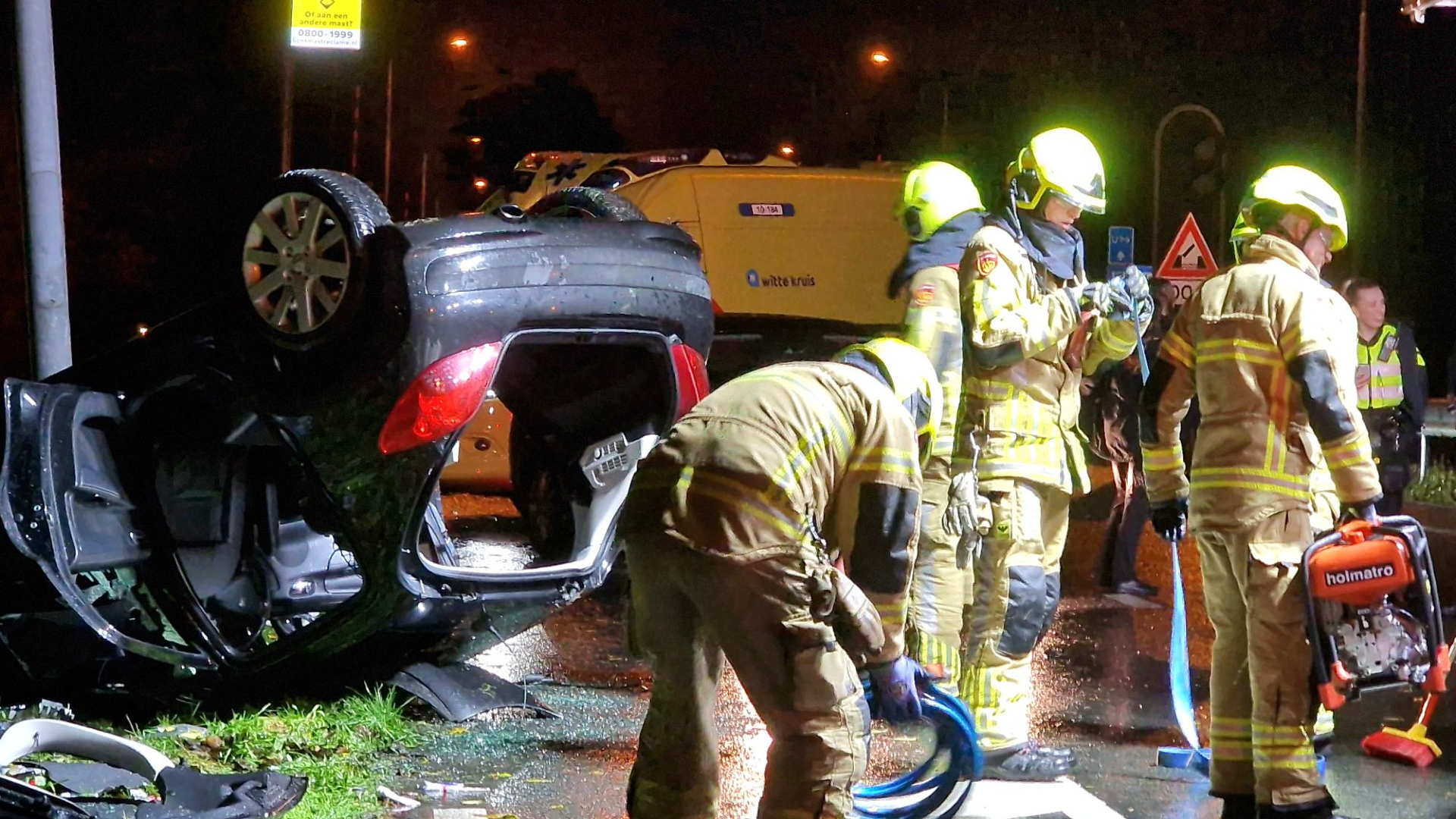 Brandweerlieden bij een auto-ongeluk met een voertuig op zijn kop, omgeven door reddingsapparatuur.