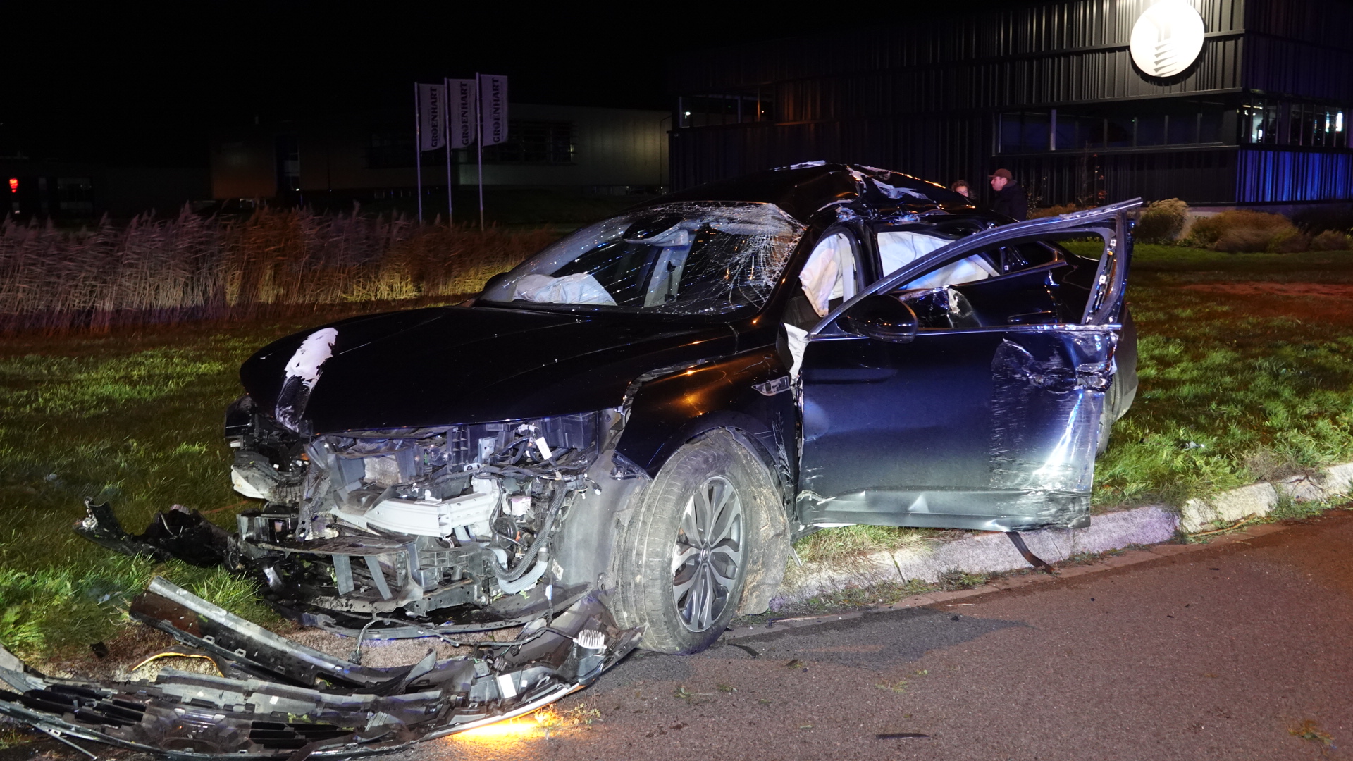 Zwaar beschadigde auto na een ongeluk, met gebroken voorruit en geopende airbags op een grasveld naast de weg.