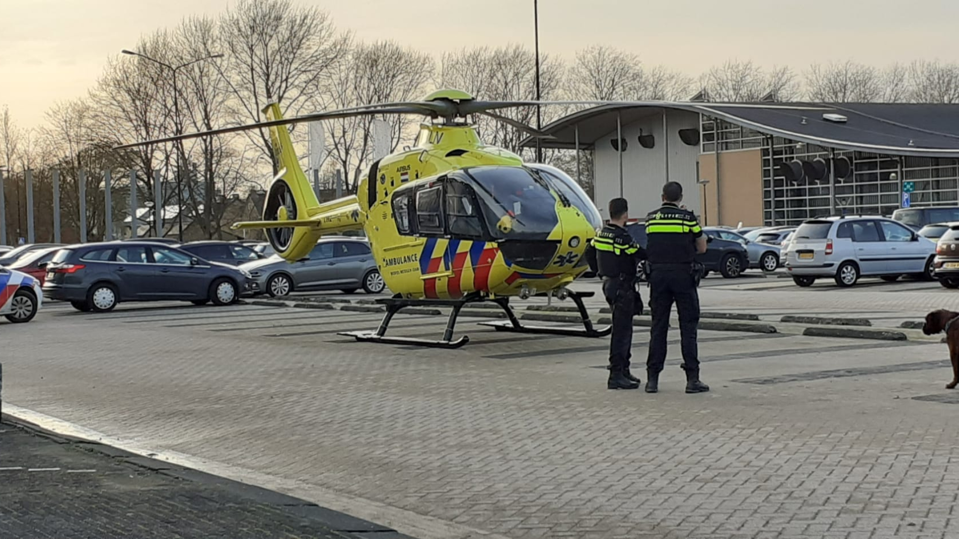 Een traumahelikopter staat geparkeerd op een parkeerplaats, met twee politieagenten en een speurhond ernaast.