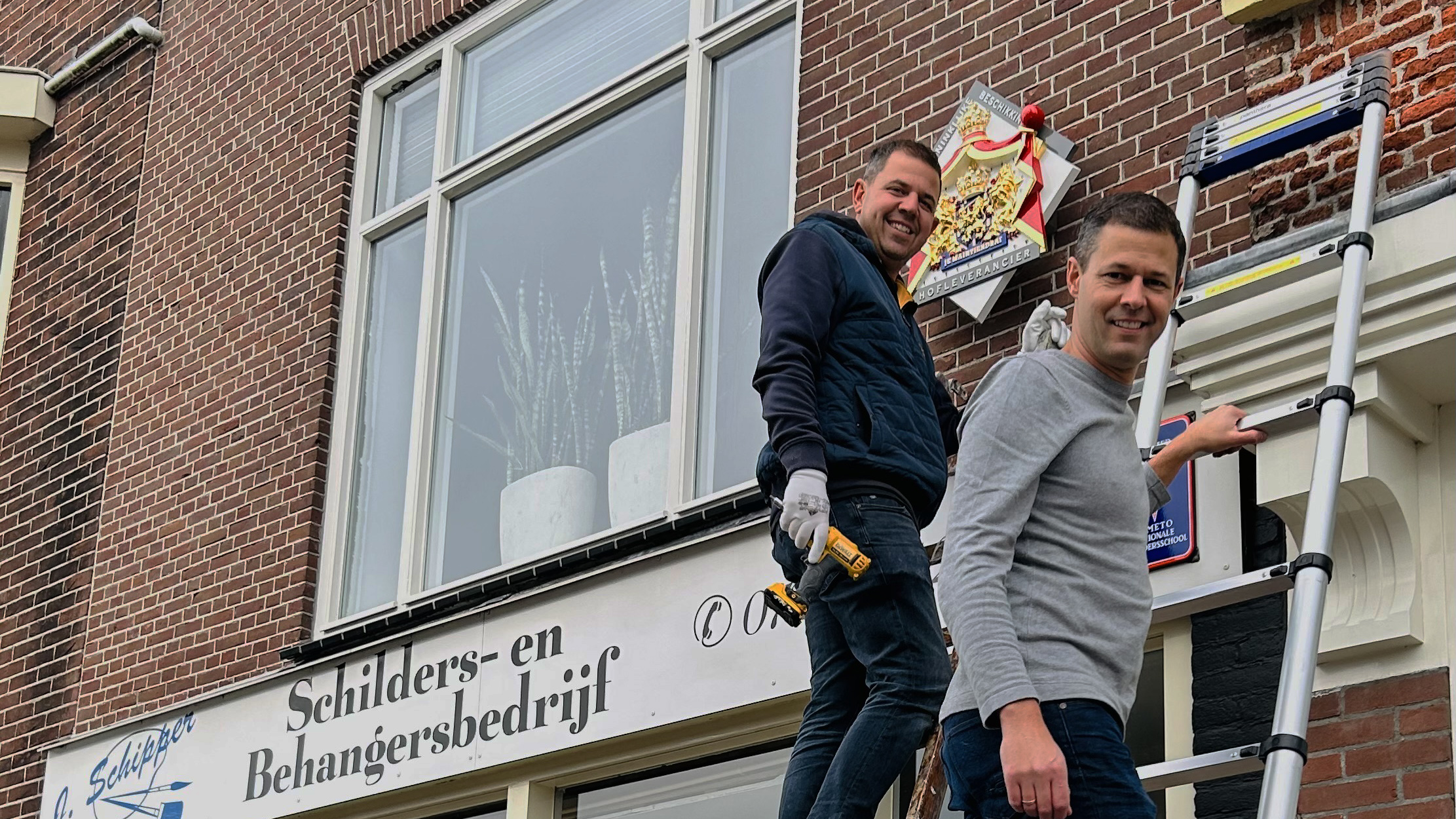 Twee mannen poseren op een ladder voor een bakstenen gebouw met een bord "Schilders- en Behangersbedrijf".