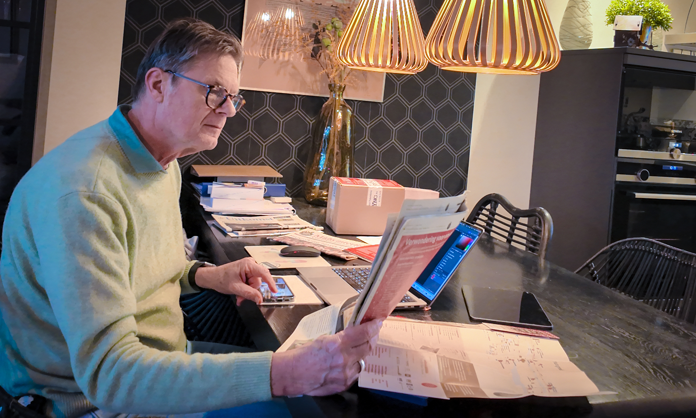 Man aan tafel leest een krant met laptop, smartphone en papieren voor zich.