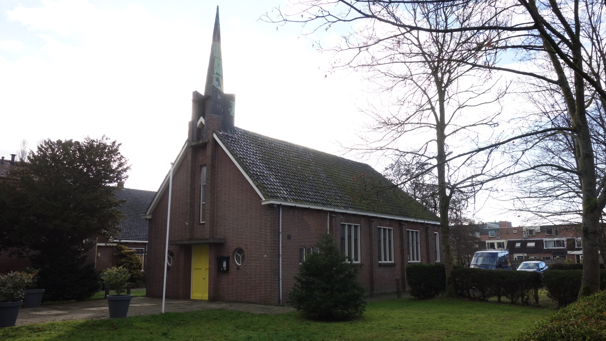 Een klein bakstenen kerkje met een groene torenspits en gele deur, omgeven door bomen en gras.