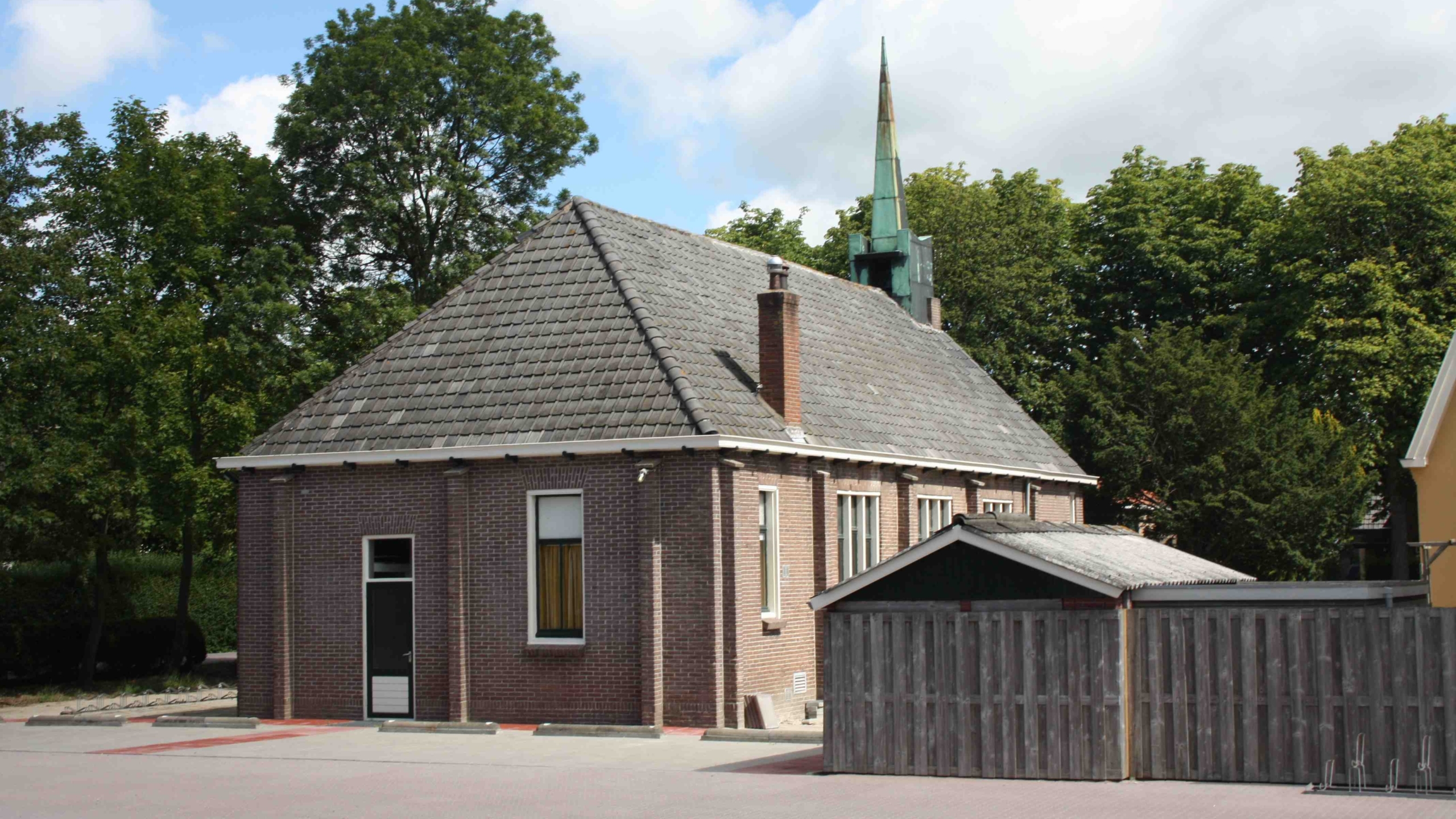 Bakstenen gebouw met grijs pannendak en groene torenspits, omgeven door bomen.