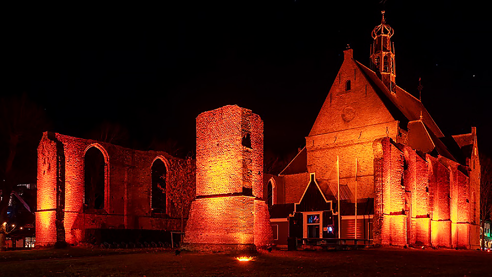 Een verlichte kerk en ruïnes bij nacht, in warme oranje tinten.