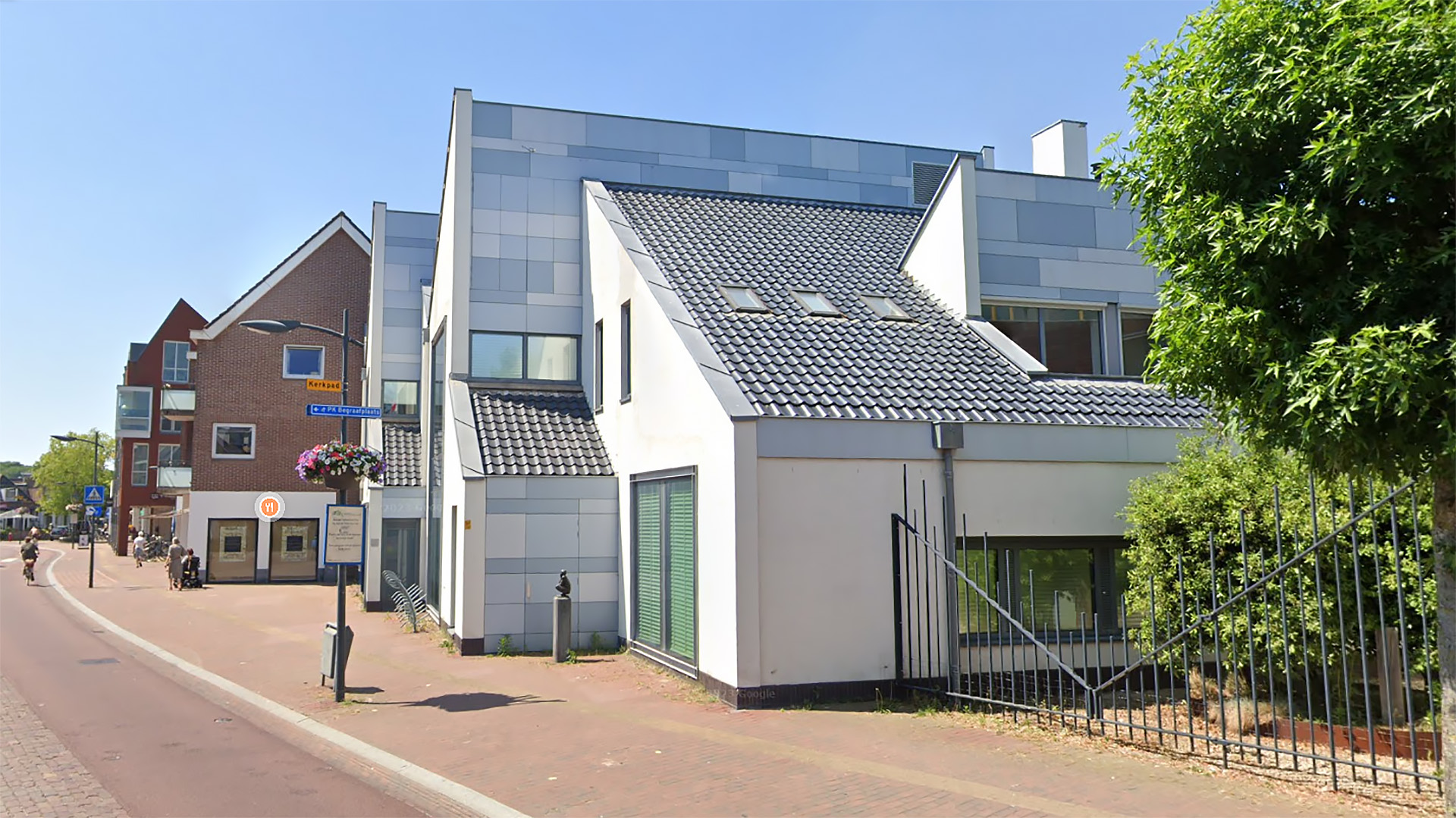 Wit gebouw met moderne architectuur, grijze dakpannen en ramen, gelegen aan een straat met een rood bakstenen huis op de achtergrond.